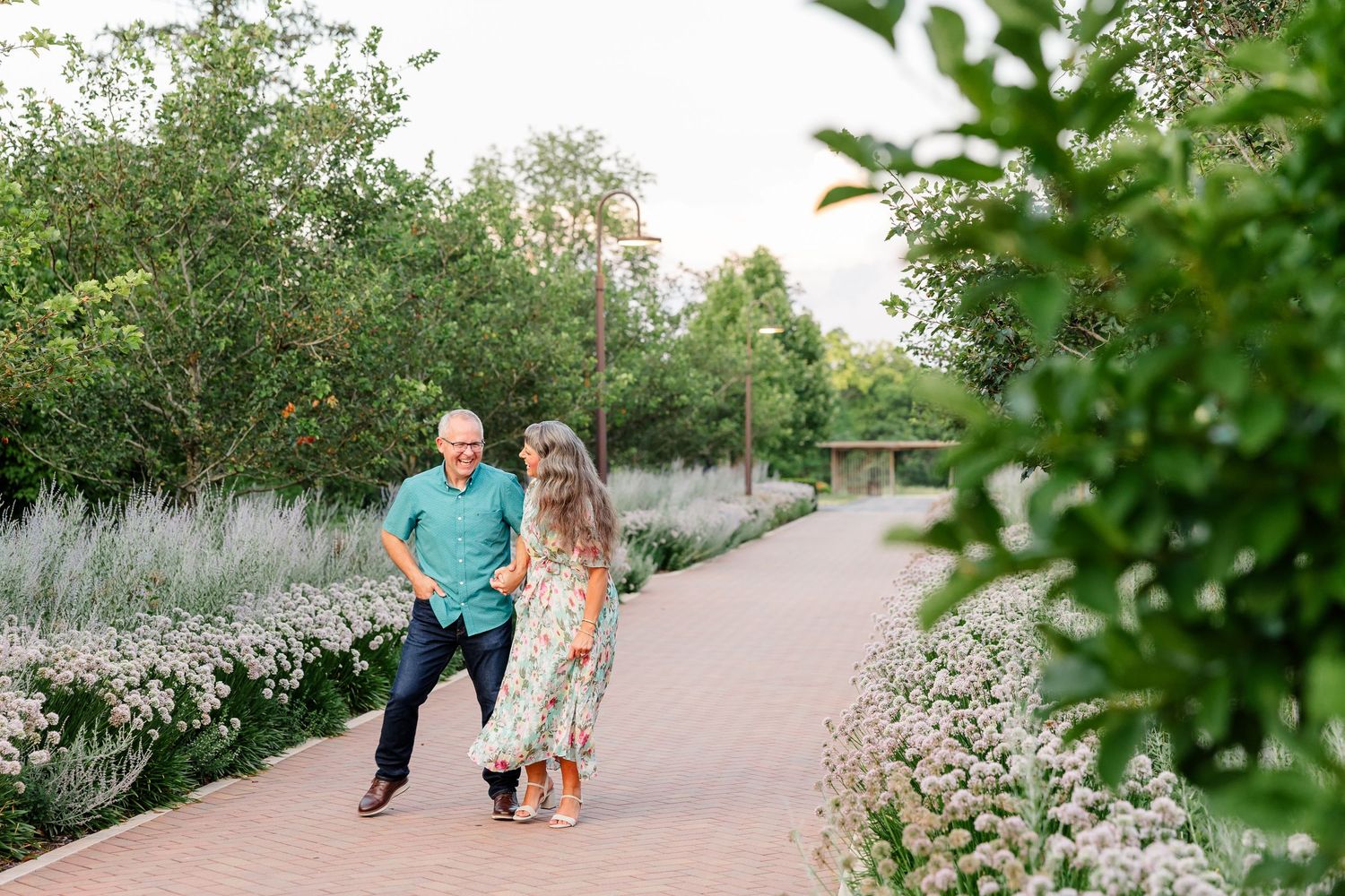 Couple walking hand in hand along a flower-lined garden path surrounded by lush green trees.