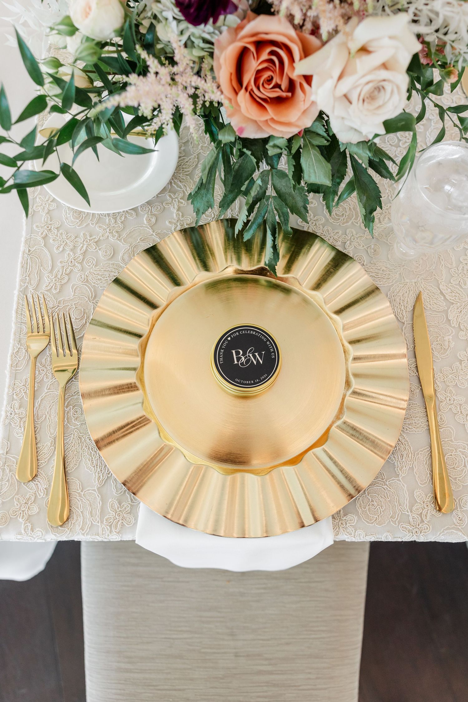 Overhead view of an elegant wedding place setting with a gold scalloped charger, gold flatware, lace linen, and peach and cream roses.