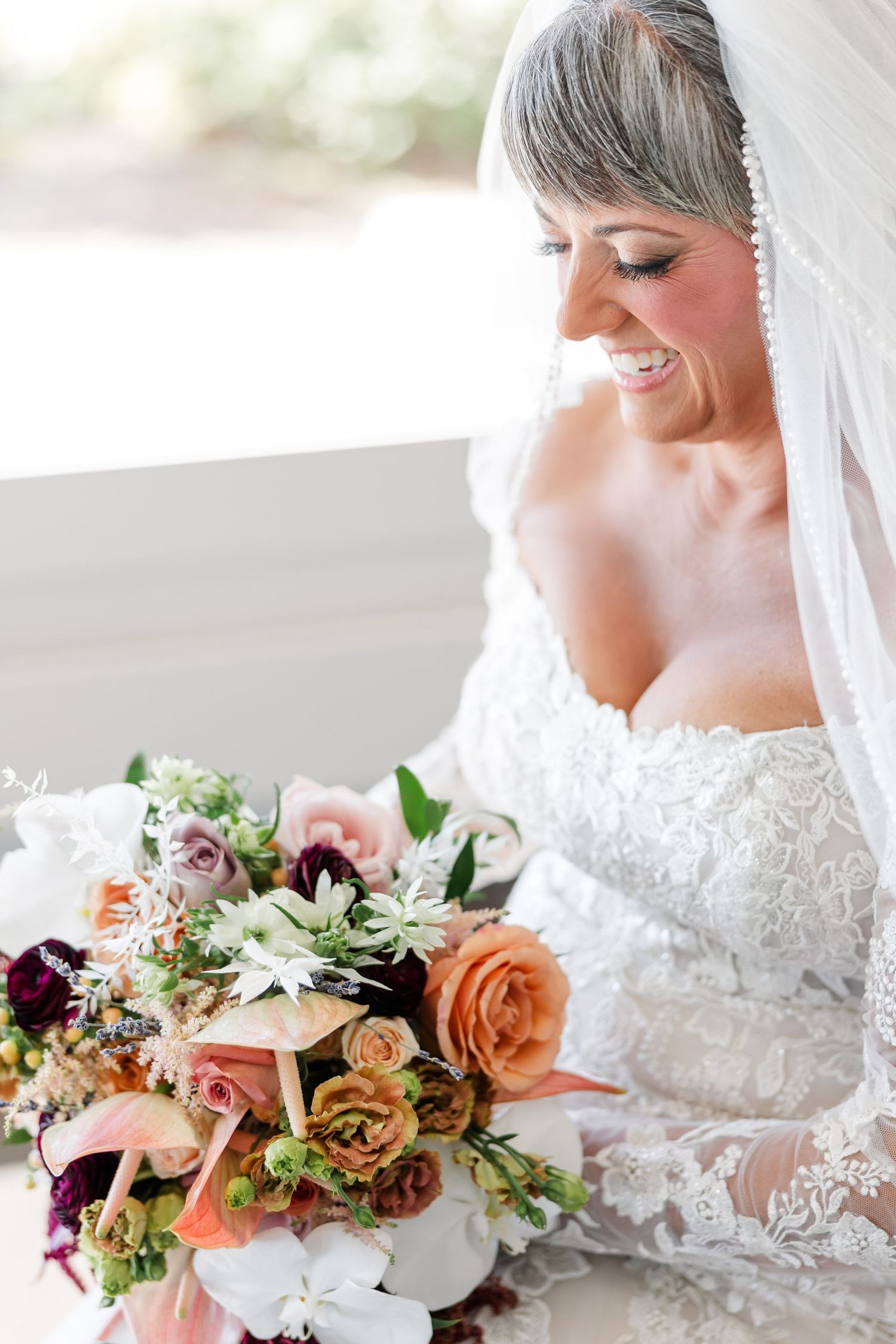 Smiling bride in a lace wedding gown and veil looks down at a colorful bouquet of peach roses, mushrooms, and white blooms.