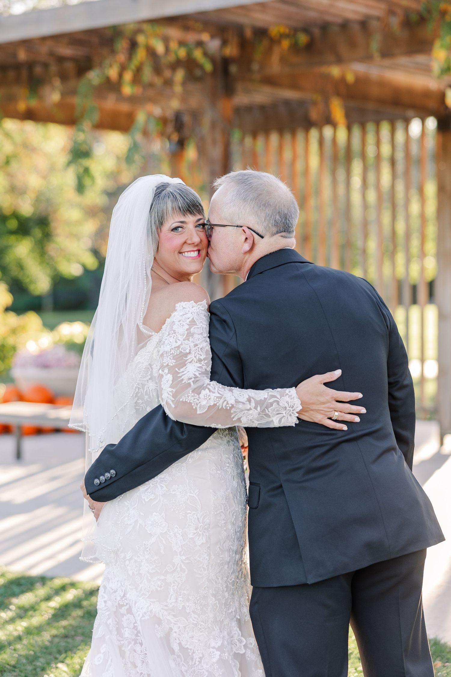 Smiling bride in a lace wedding gown and veil is kissed on the cheek by her groom in a dark suit under a wooden pergola.