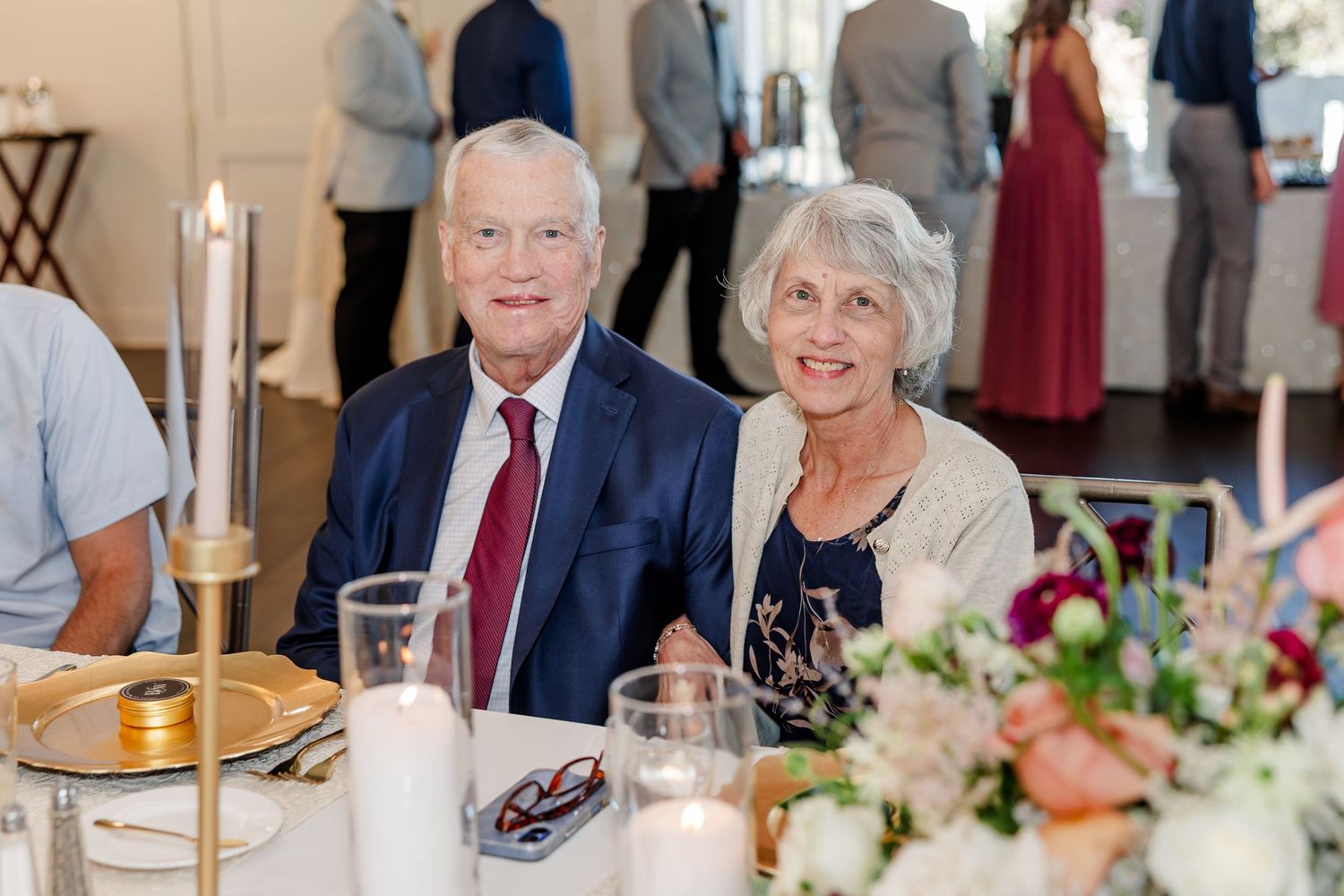 Smiling elderly couple seated at a wedding reception table with candles and a floral centerpiece while guests mingle behind them.