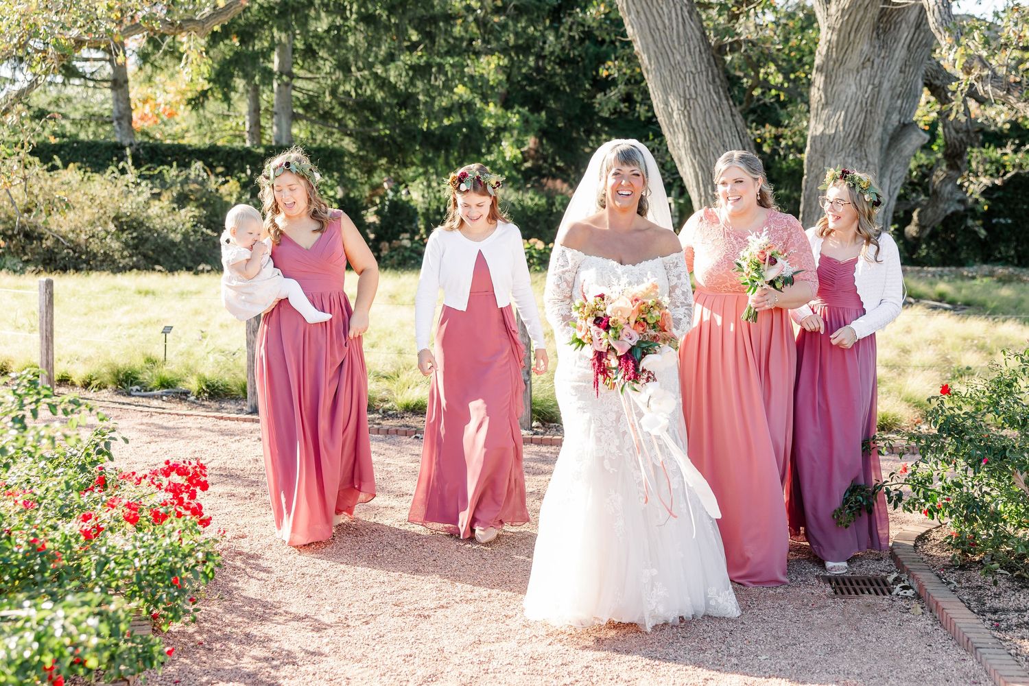 Bride holding a bouquet walks along a garden path with bridesmaids in rose and mauve dresses, one carrying a baby, on a sunny day.