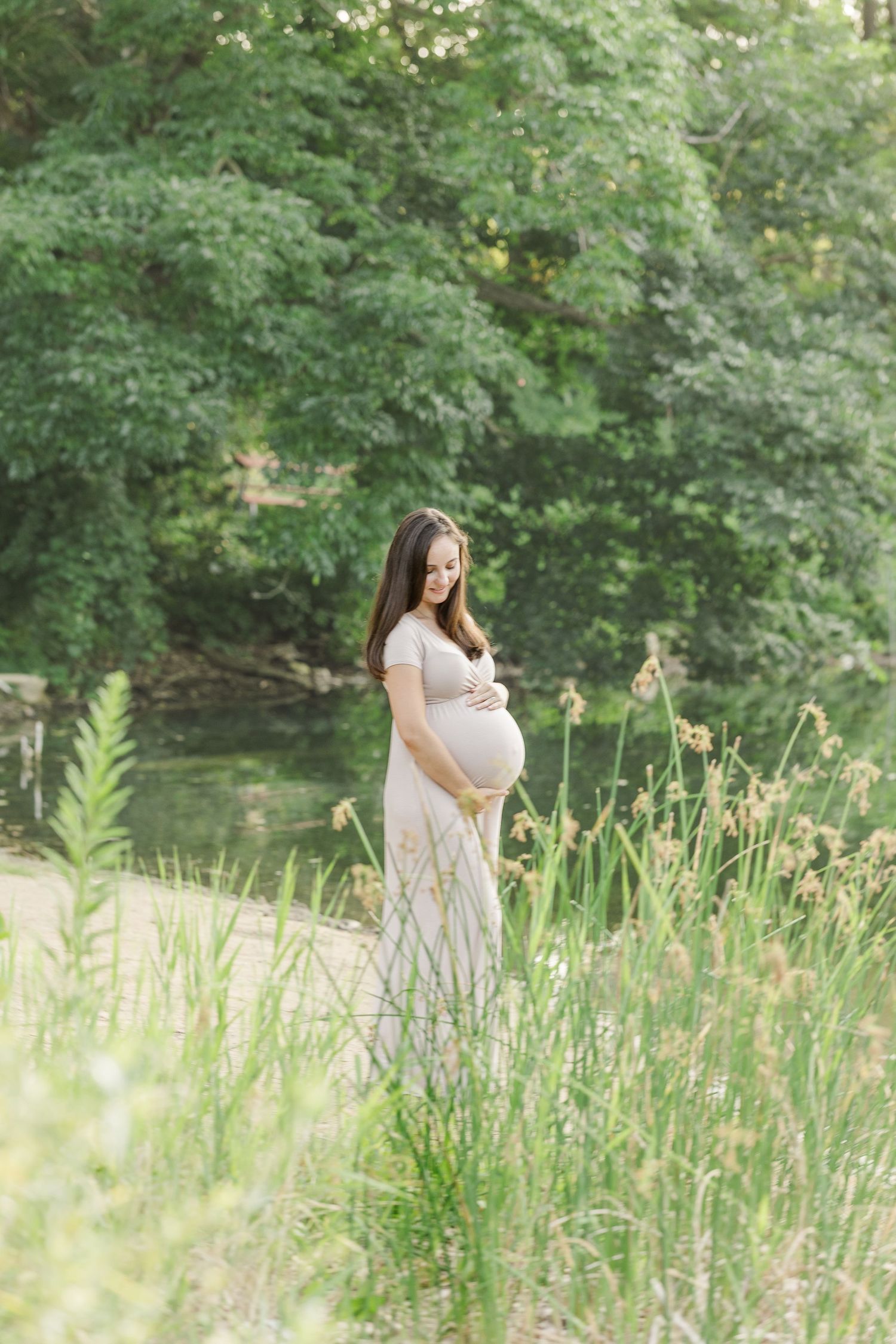 A woman in a white dress stands in tall grass by a river, cradling her pregnant belly in a tranquil natural setting.