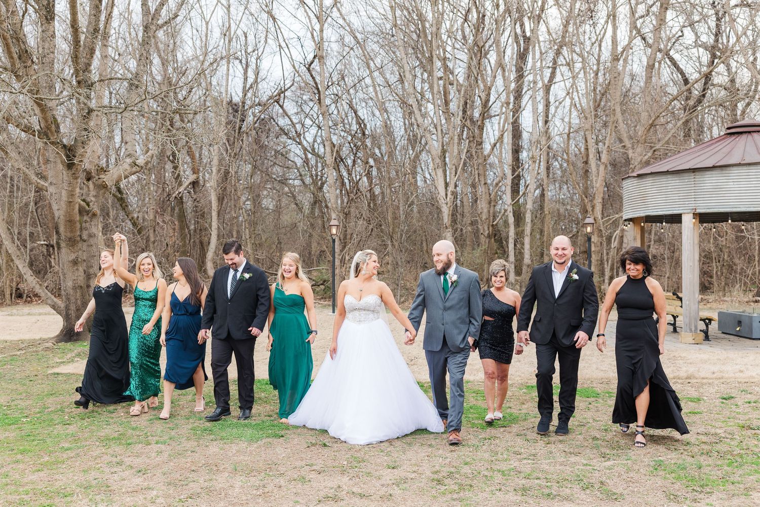 Wedding party walking outdoors in green and black attire among bare winter trees.