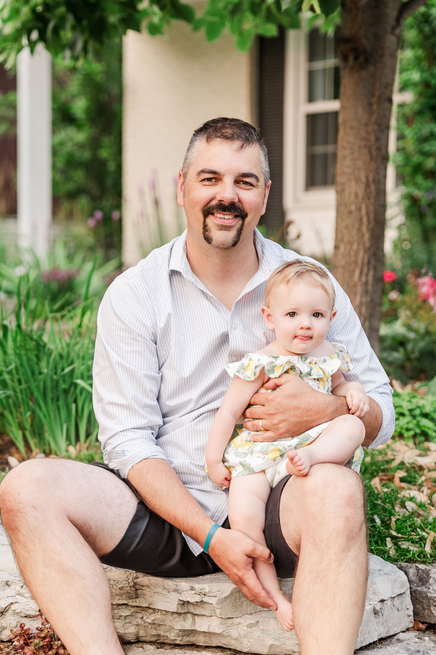 Portrait in a garden setting featuring baby in floral outfit sitting with adult on stone ledge.