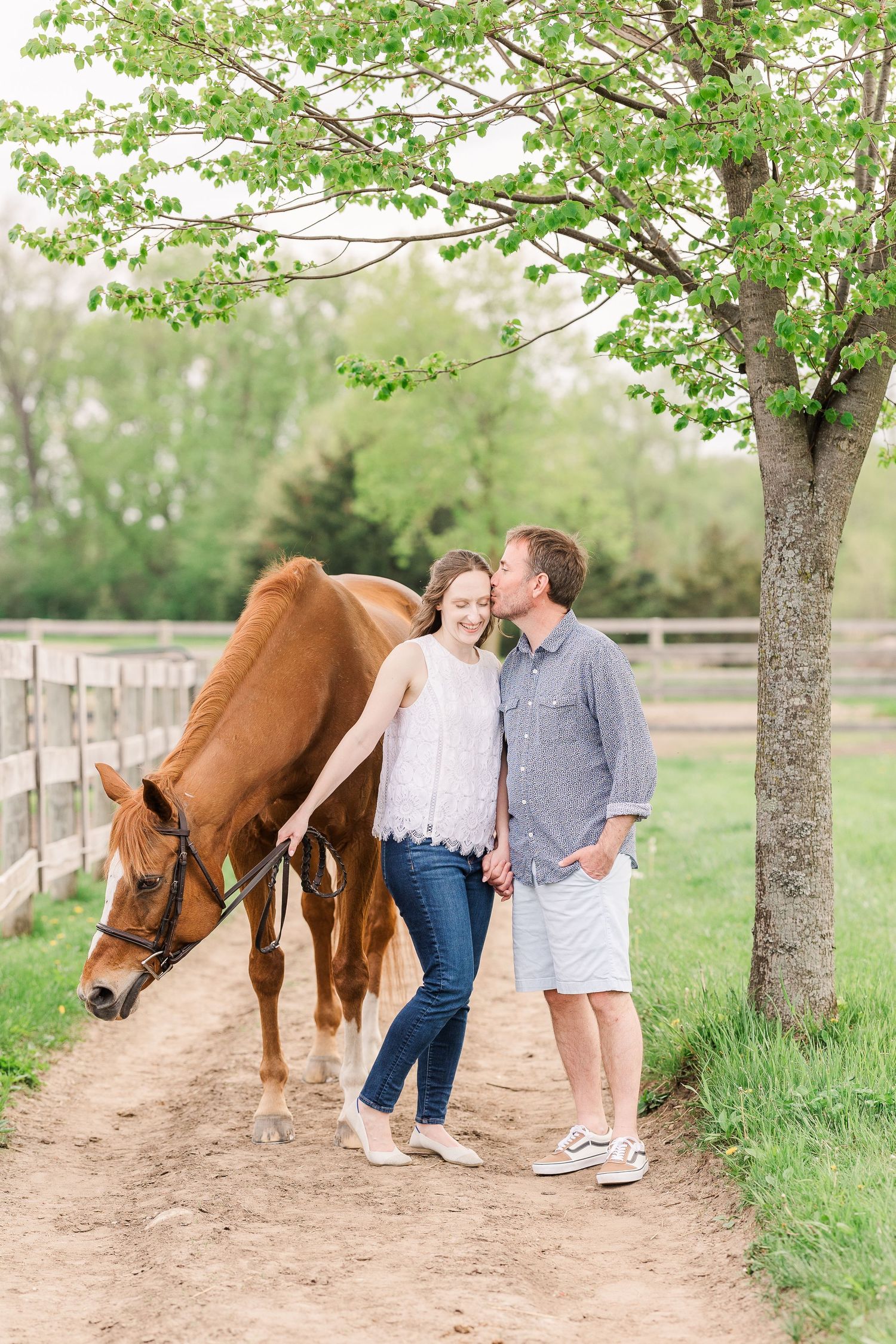 A couple stands with a chestnut horse on a dirt path under a blooming tree near a wooden fence.