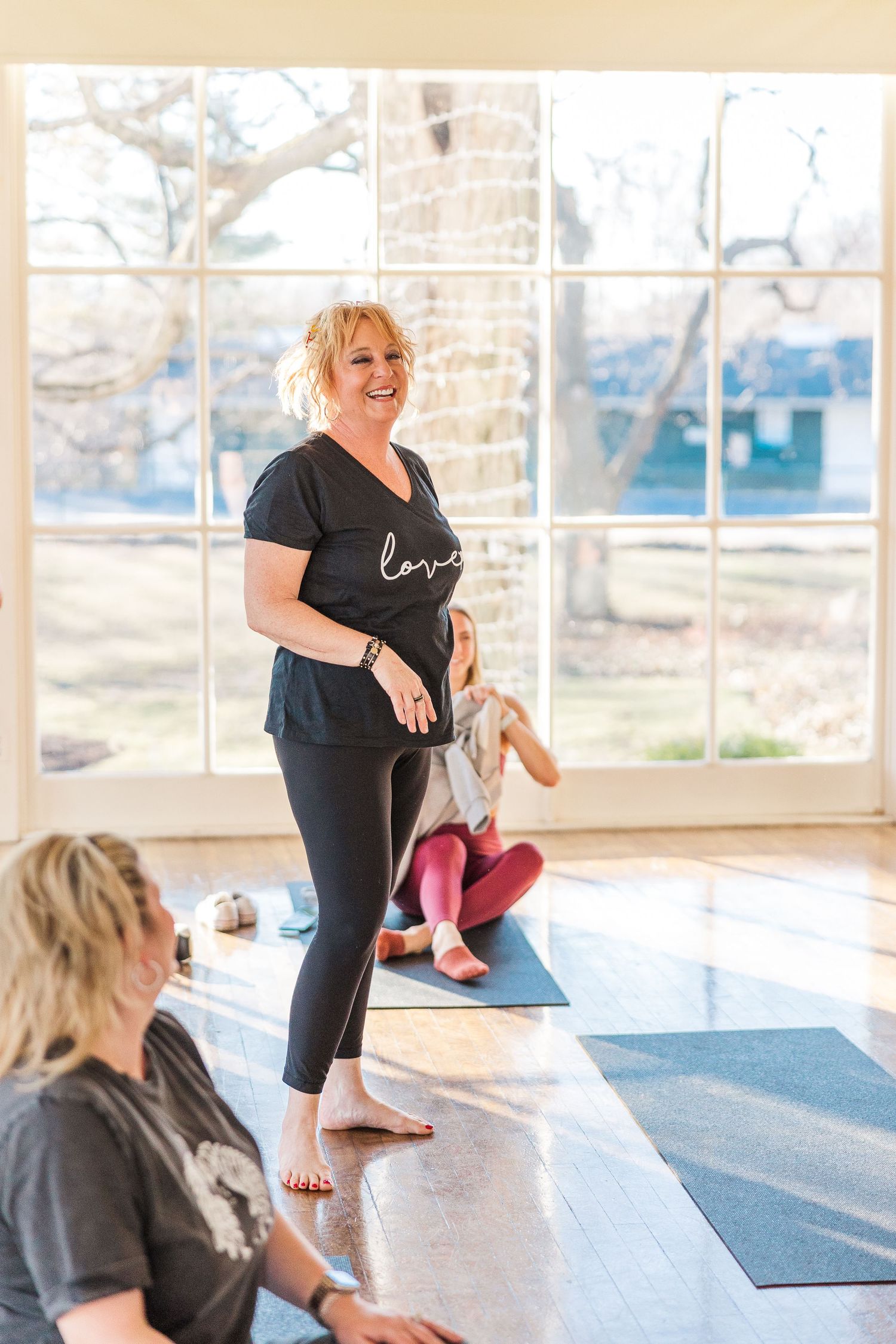 A yoga instructor demonstrates poses on a geometric patterned mat in front of large windows.