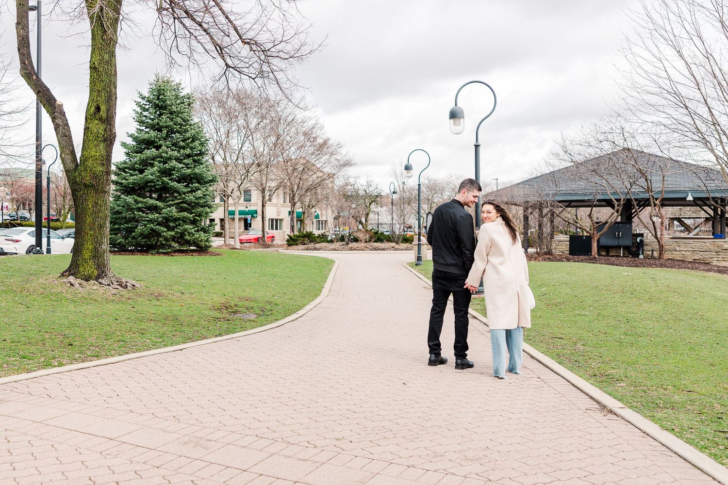 Two people stroll down a curved brick pathway near a gazebo and evergreen trees in a park setting.