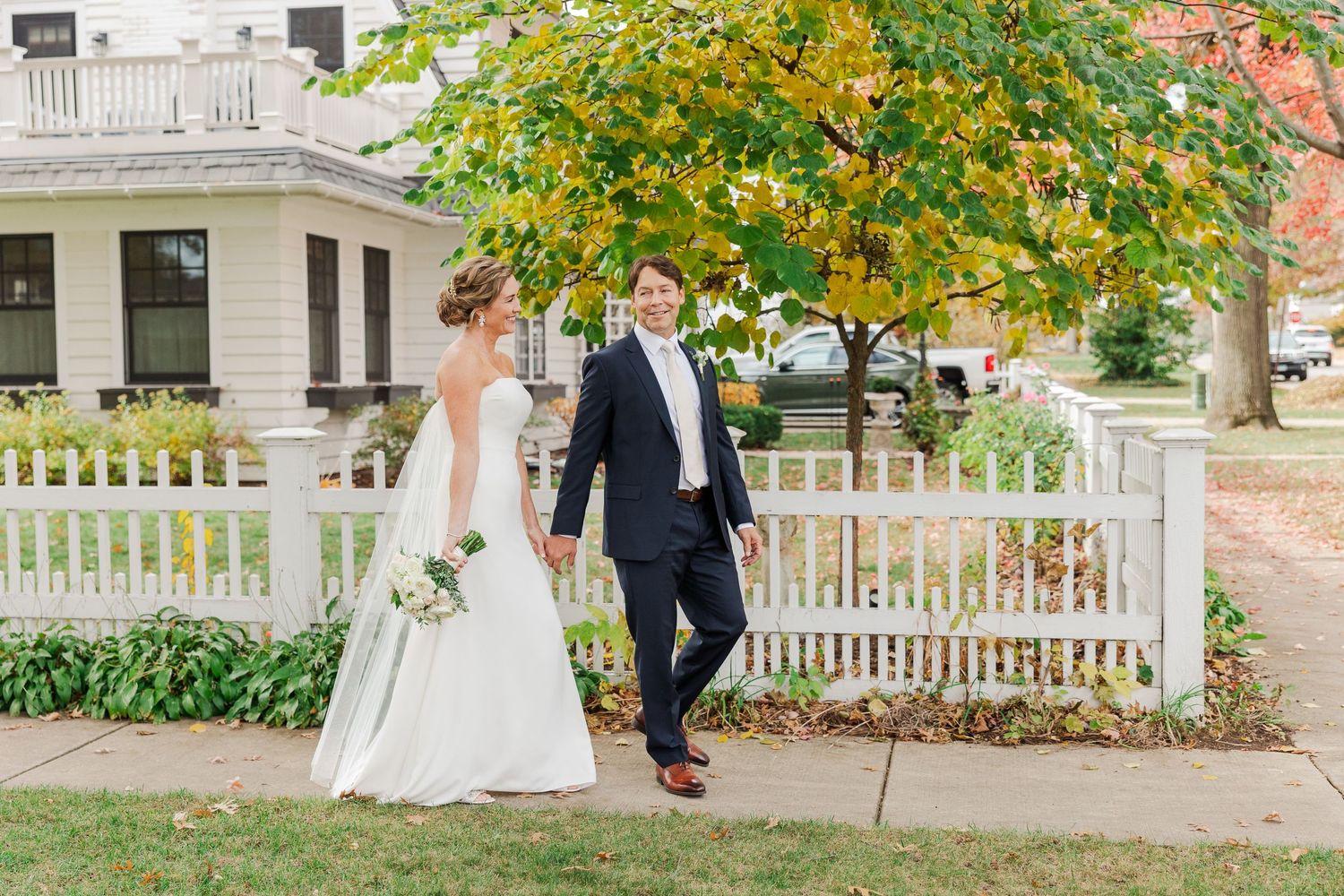 Newlyweds stand by white fence with autumn foliage and white house in background.