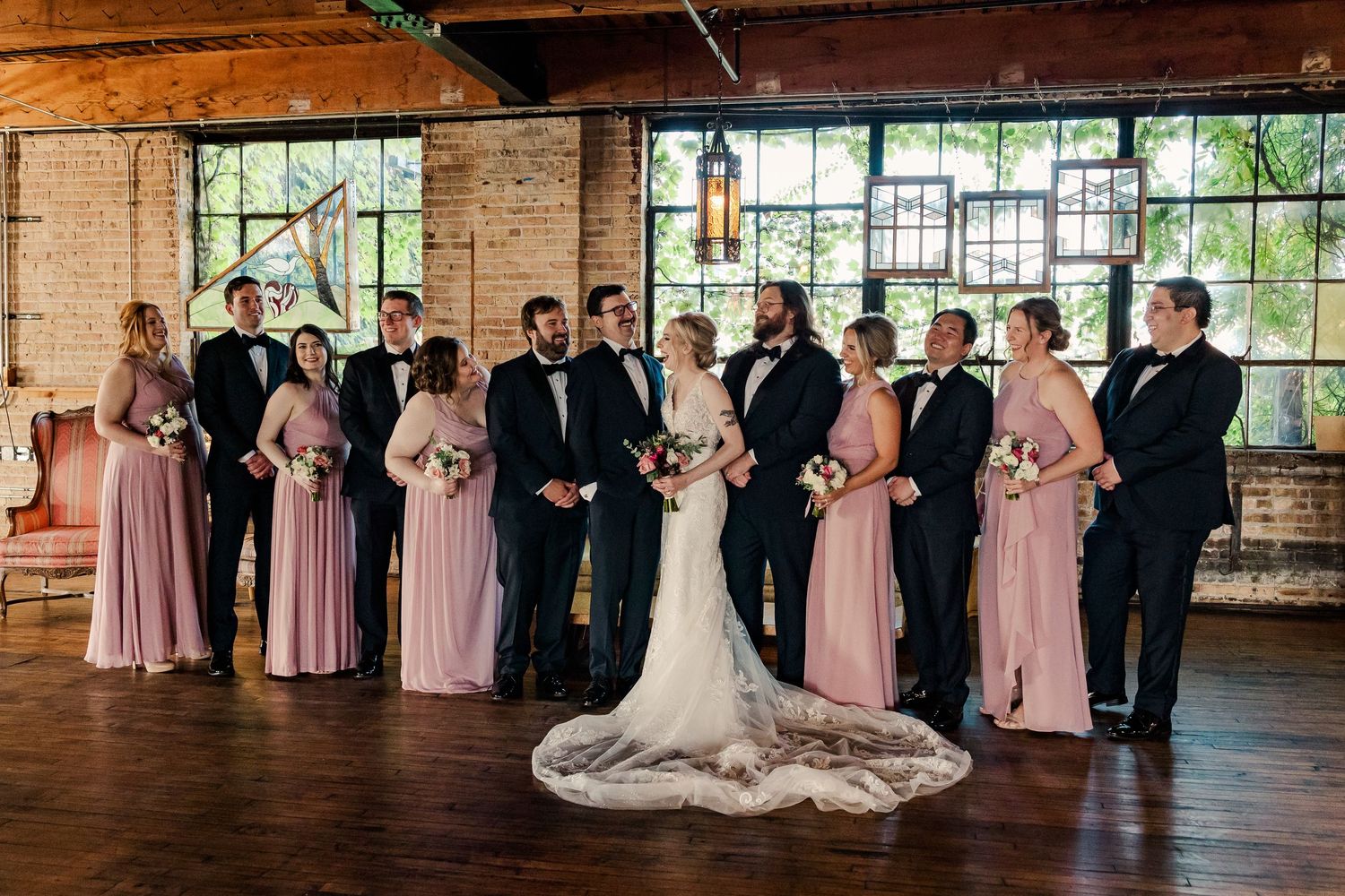 Wedding party poses in symmetrical formation with pink bridesmaid dresses and black tuxedos.