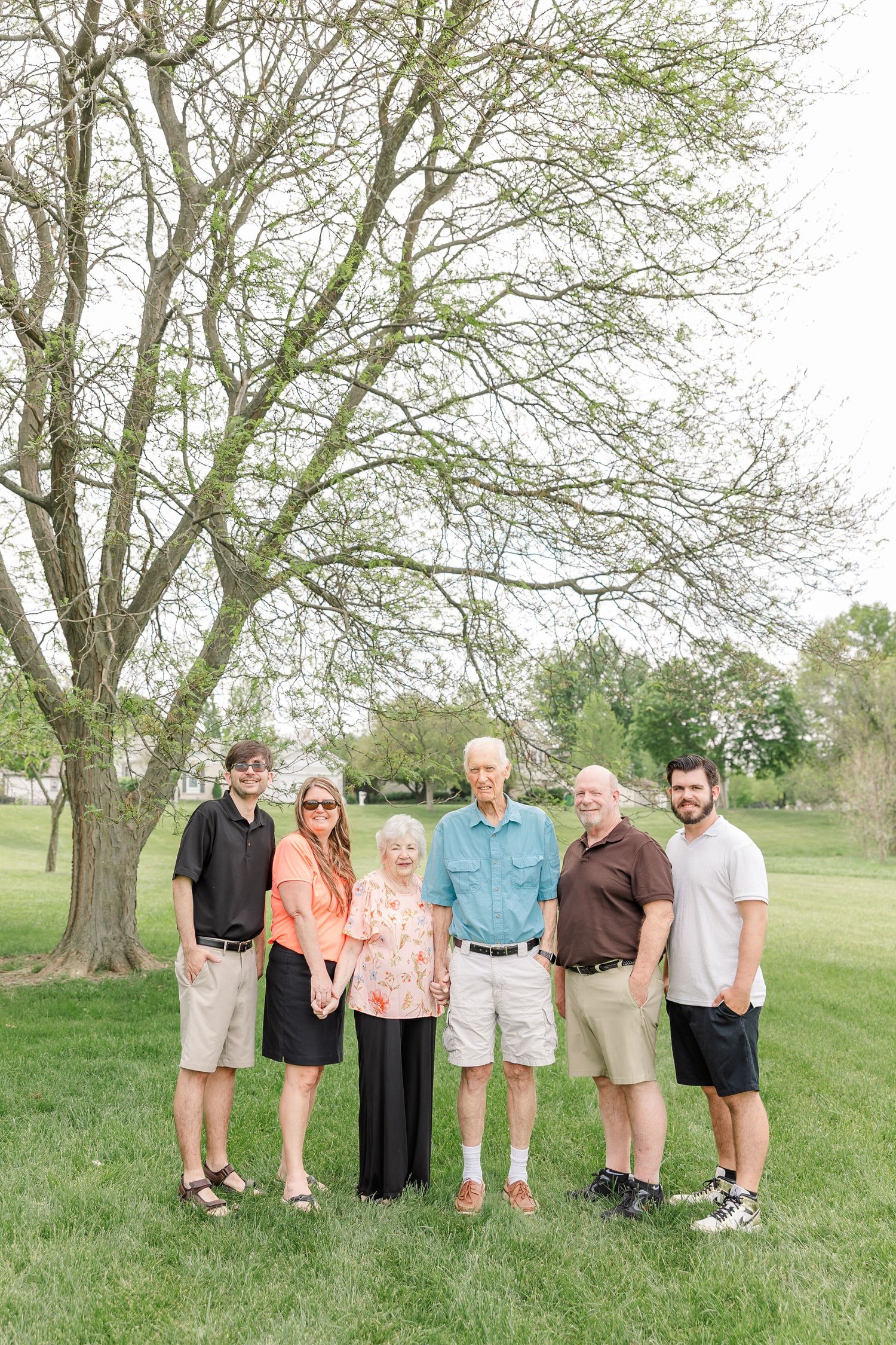 Multi-generational family of six poses together outdoors on green grass beneath a large tree on a spring day.