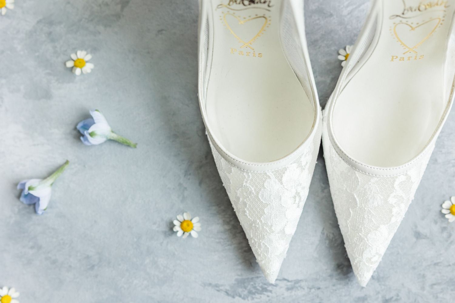 White lace bridal heels surrounded by delicate daisies and blue flowers on a gray marble surface.