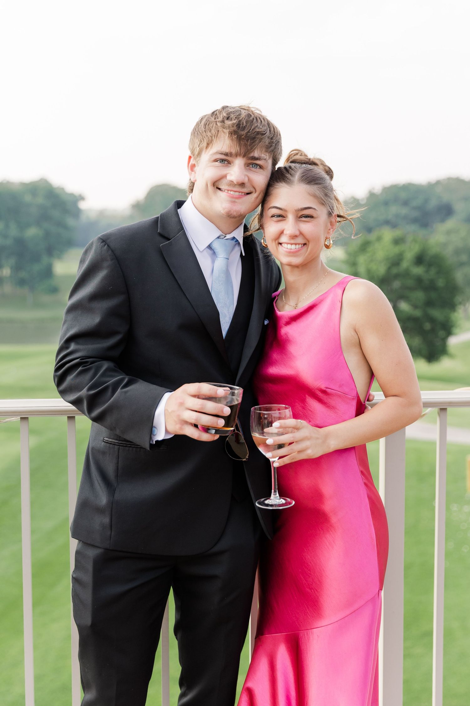 Smiling couple in formal attire holds wine glasses on an outdoor balcony overlooking a green golf course.