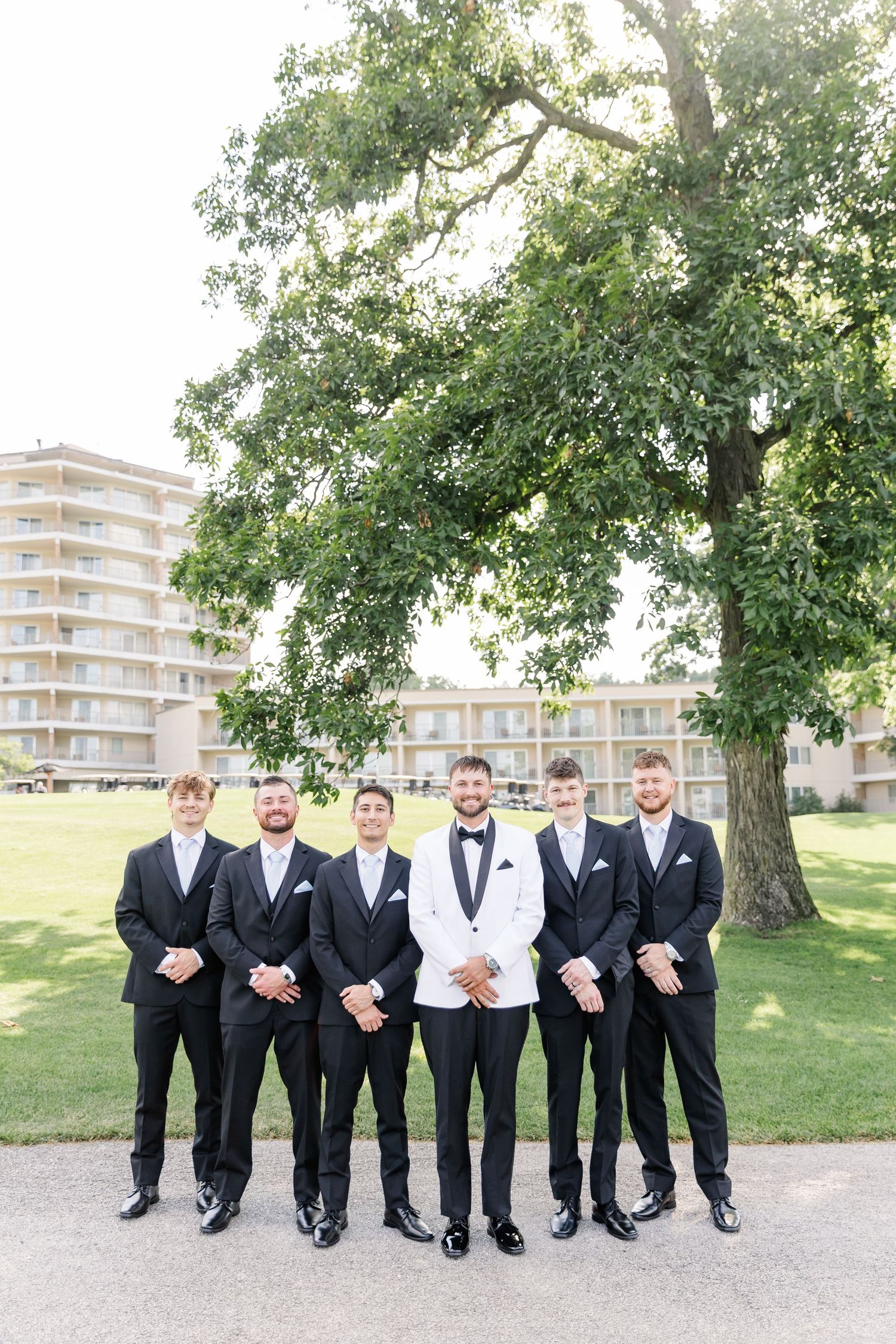 Groom in white tuxedo jacket poses with six groomsmen in black suits outdoors near a large tree.