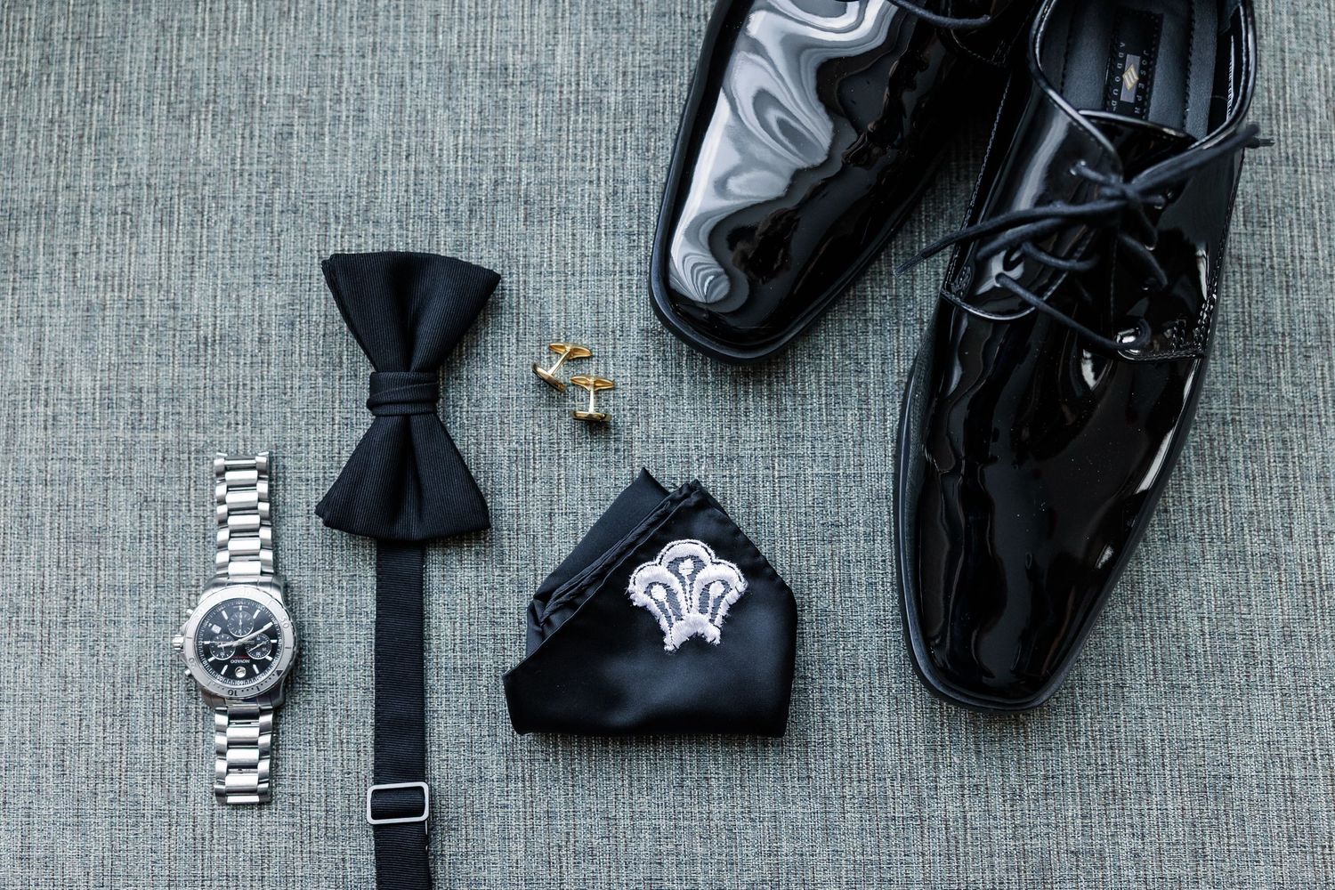 Groom accessories flat lay with black bow tie, silver watch, gold cufflinks, pocket square, and patent leather dress shoes.