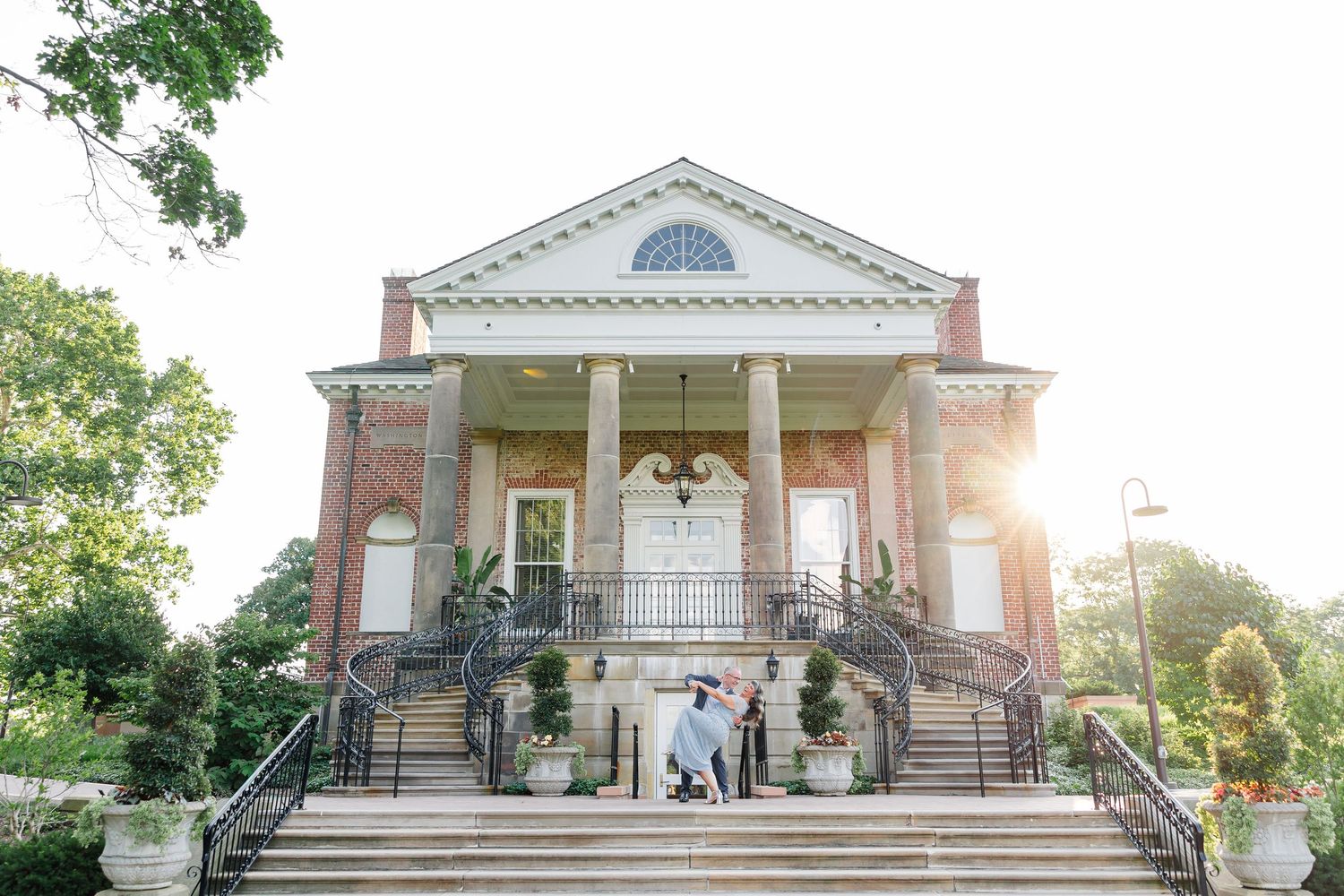Couple embracing on steps of elegant brick colonial wedding venue with white columns and lush greenery.