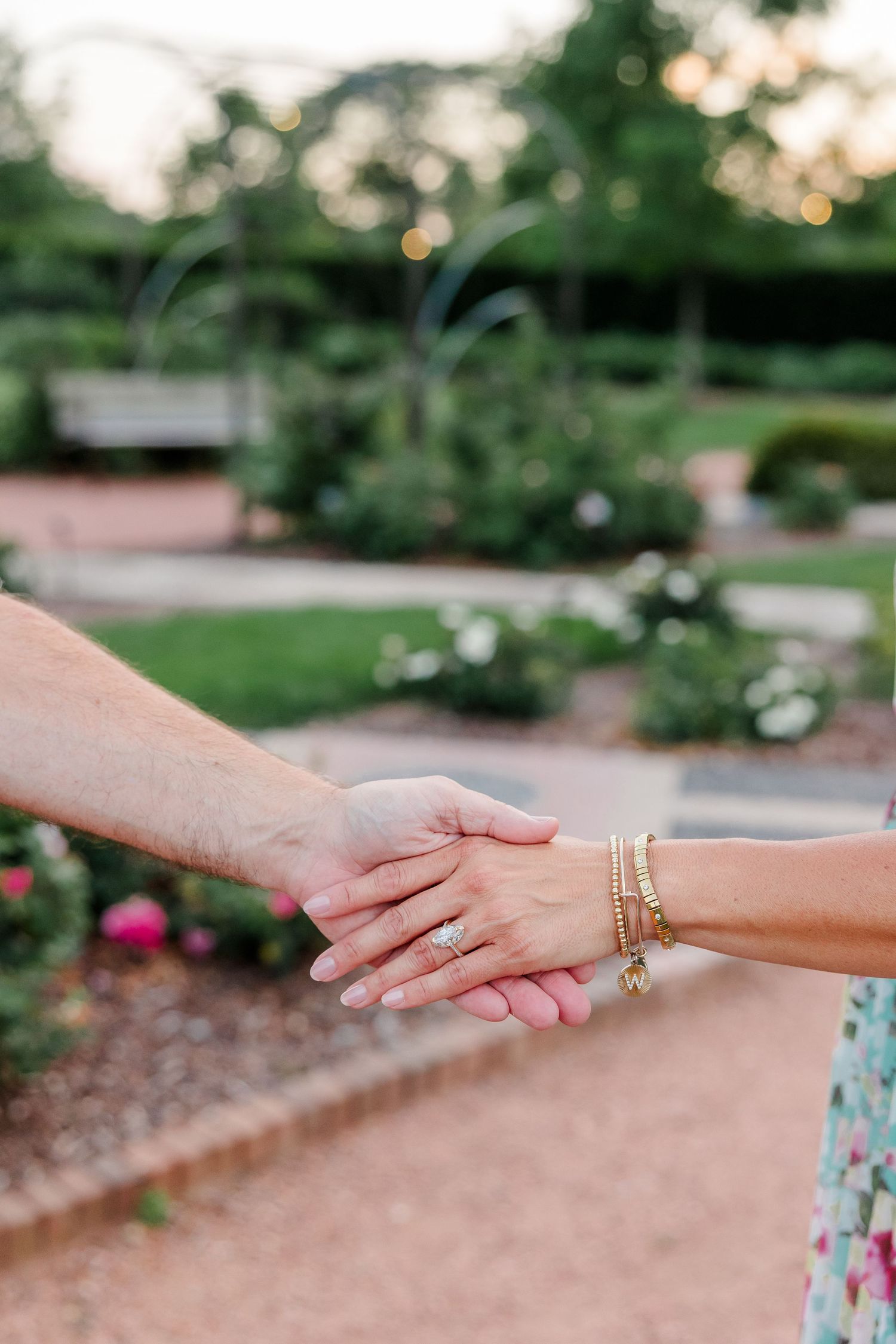 Close-up of couple holding hands showing engagement ring and gold bracelets in rose garden.