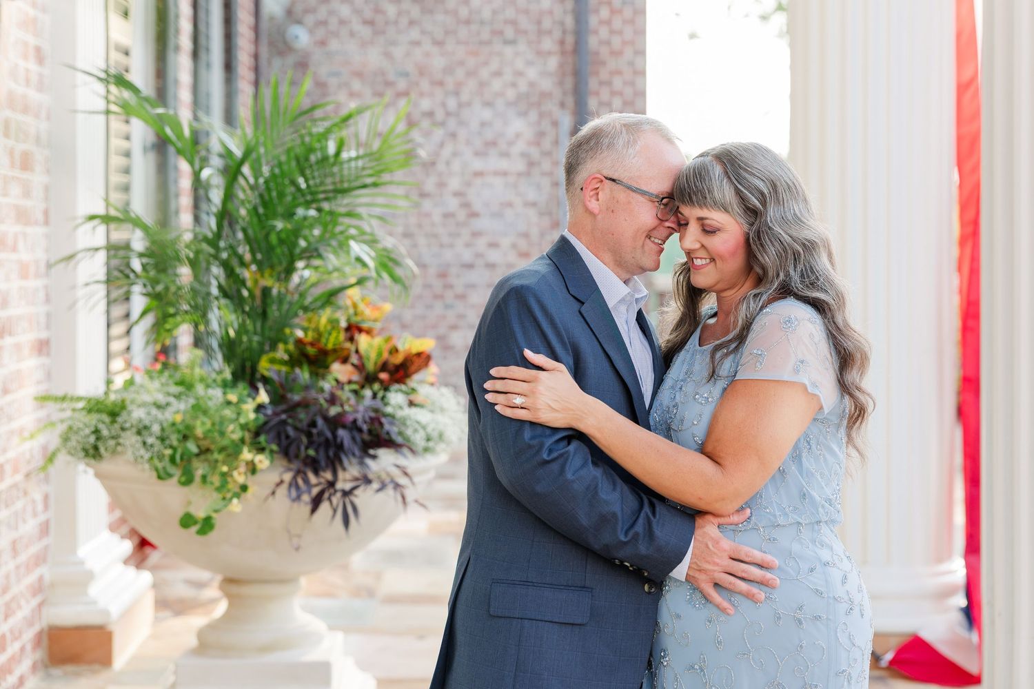 An older couple embraces and smiles at each other outdoors near a brick building with tropical floral arrangement.