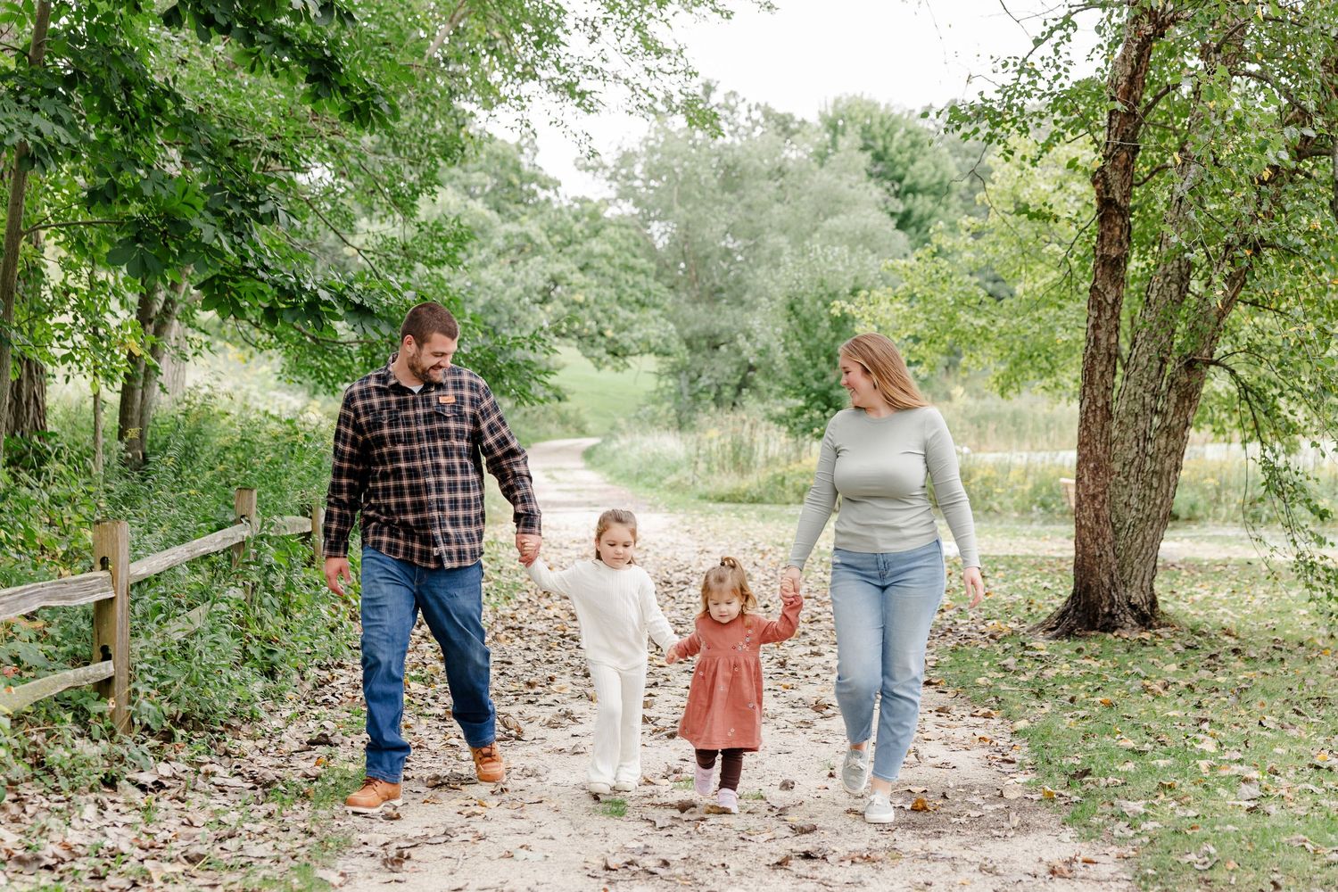 Family of four walking hand-in-hand on a scenic dirt path surrounded by lush green trees.