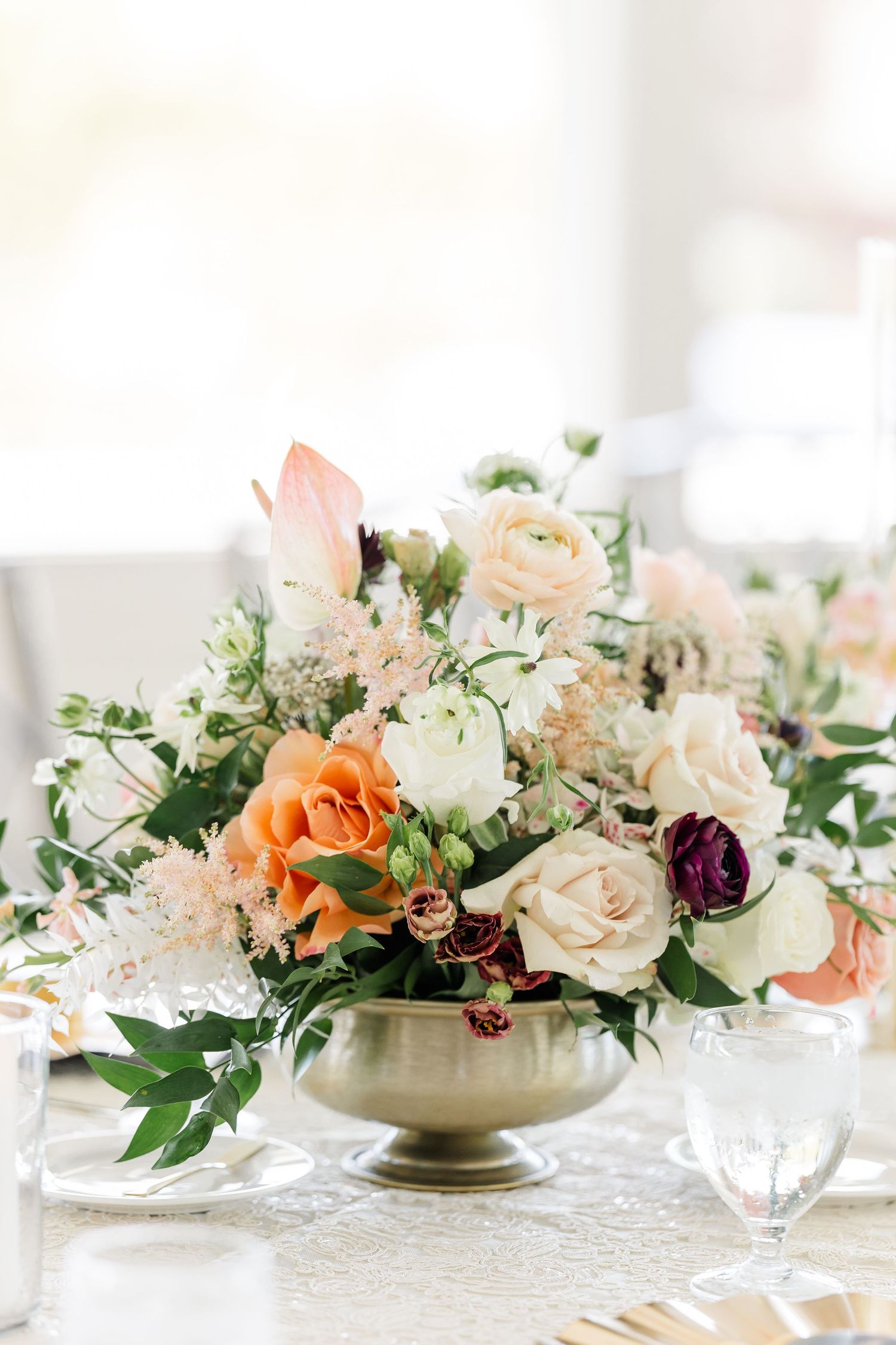 Romantic wedding centerpiece of peach, blush, and burgundy roses with greenery in a gold pedestal vase on an elegantly set reception table.