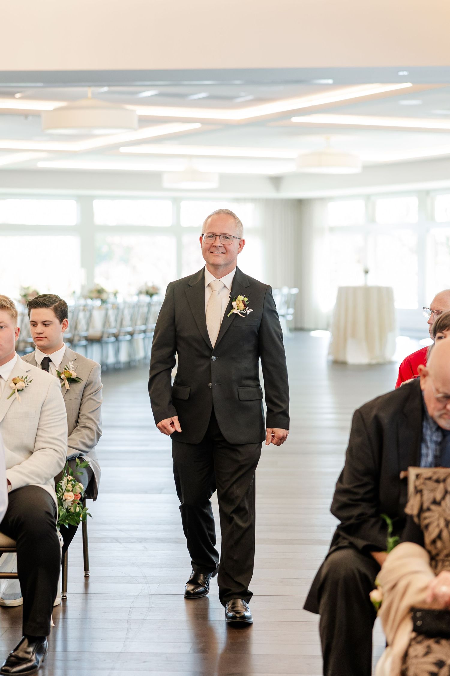 A smiling man in a dark suit with a boutonniere walks down the aisle at a bright indoor wedding ceremony as guests look on.