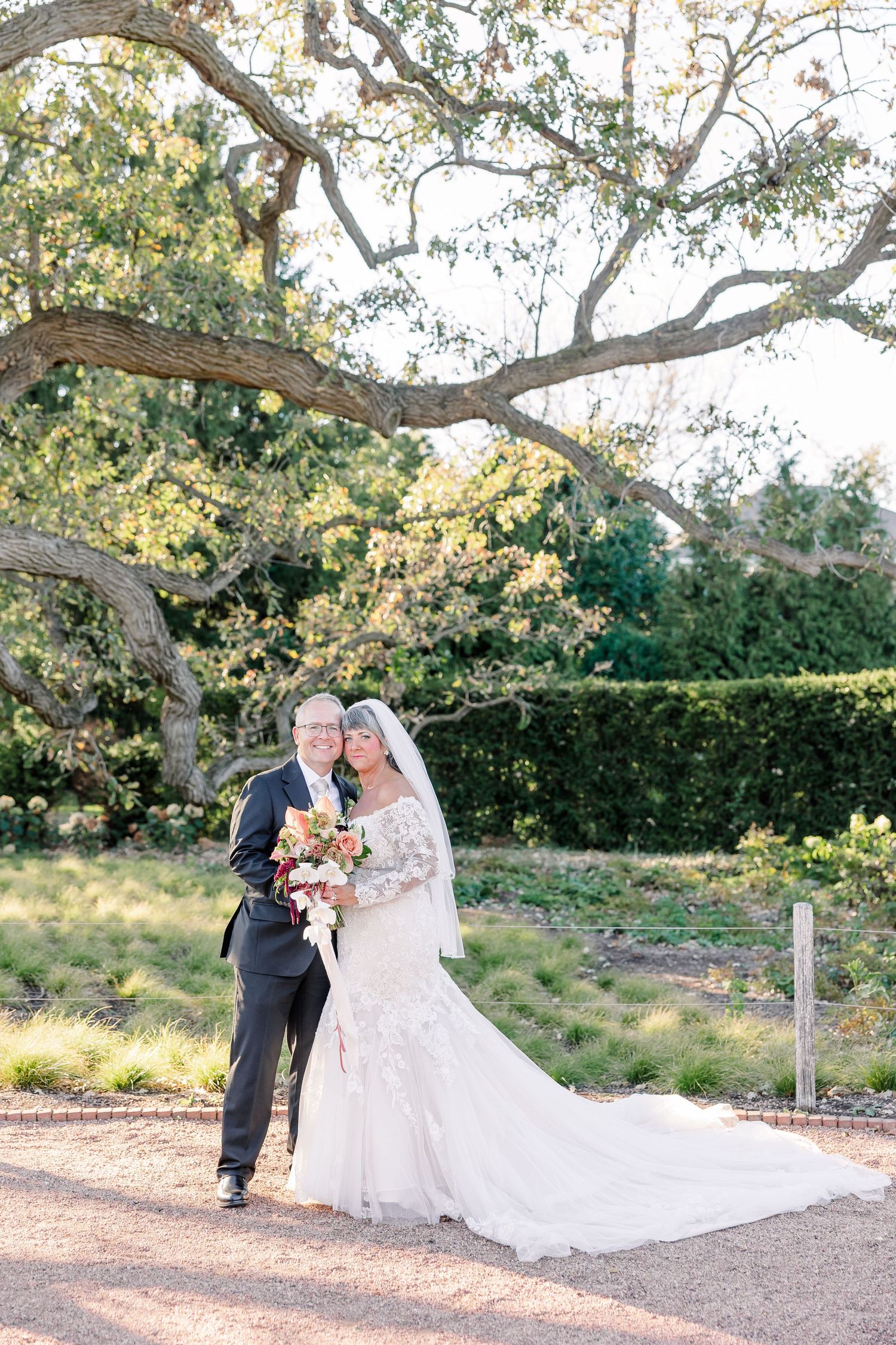 Smiling bride in a lace gown and veil holding a bouquet embraces her groom in a dark suit beneath a sprawling oak tree in a garden.