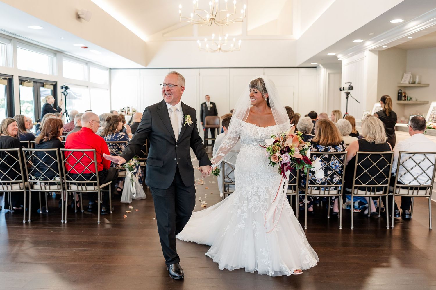 Smiling bride in a white mermaid gown holding a bouquet walks hand in hand with a groom in a dark suit past seated wedding guests.