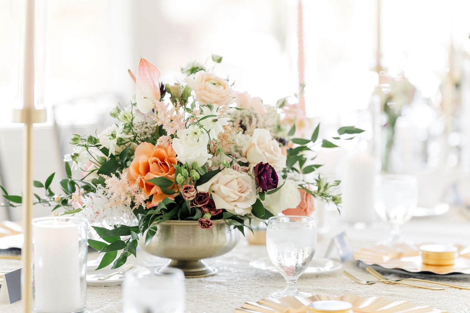 Elegant wedding reception table centerpiece with blush, peach, and white roses in a gold pedestal vase beside candles and glassware.
