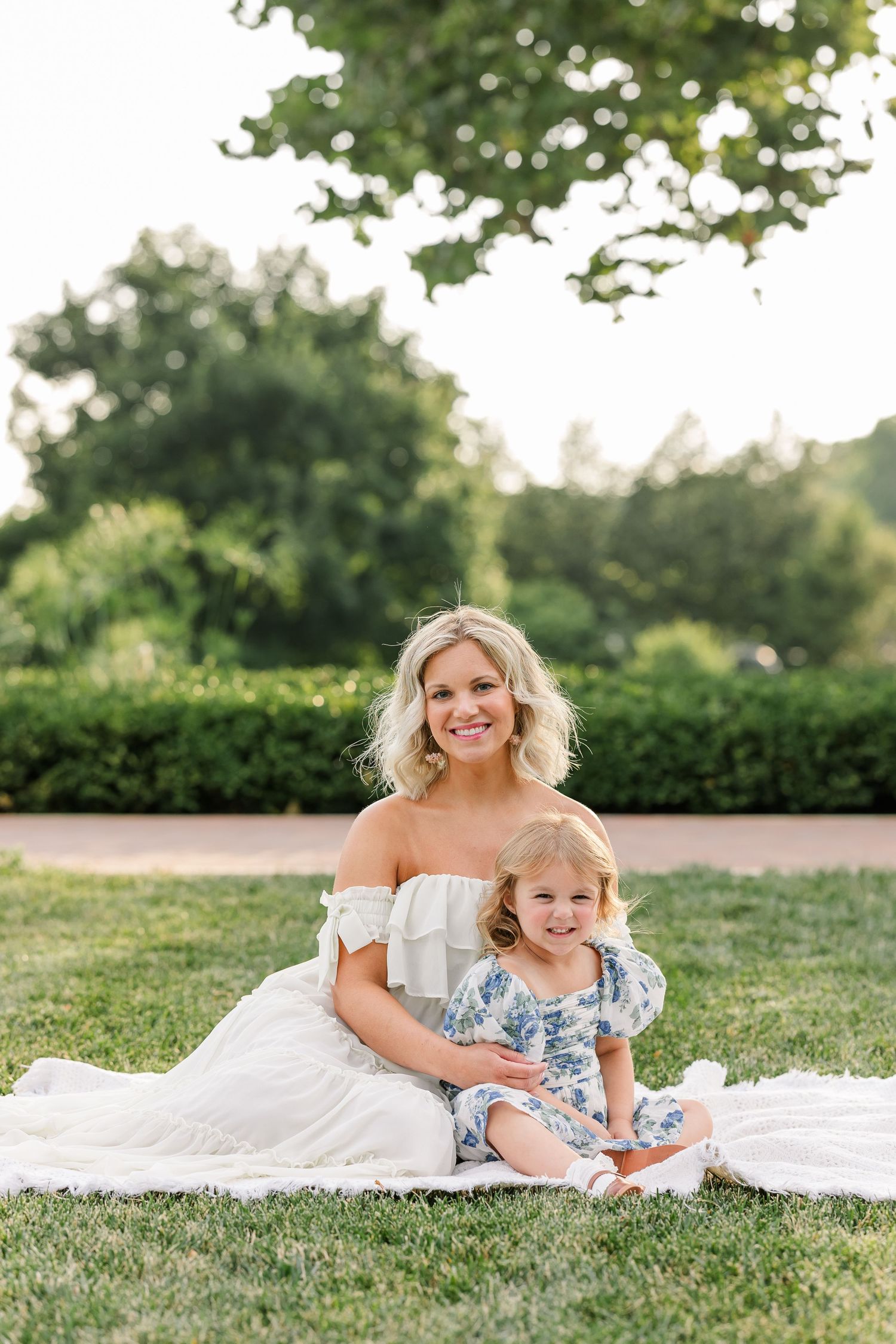 Smiling blonde mother in white off-shoulder dress sitting on blanket in park with young daughter in floral outfit.