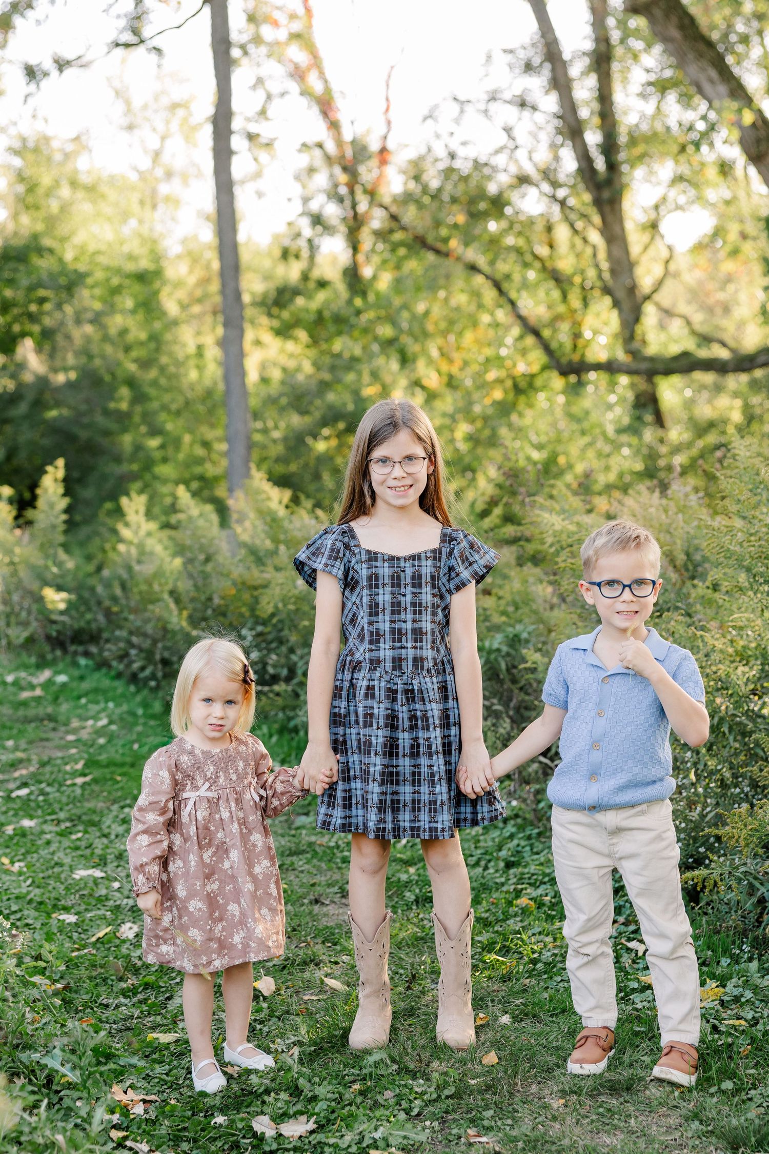 Three siblings holding hands beneath backyard trees during fall portraits in St. Charles, IL.