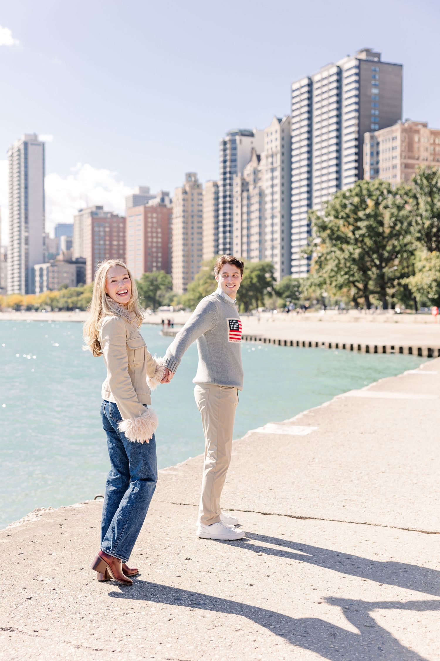 Couple holding hands and walking along Chicago lakefront path with city skyline and turquoise water in background.