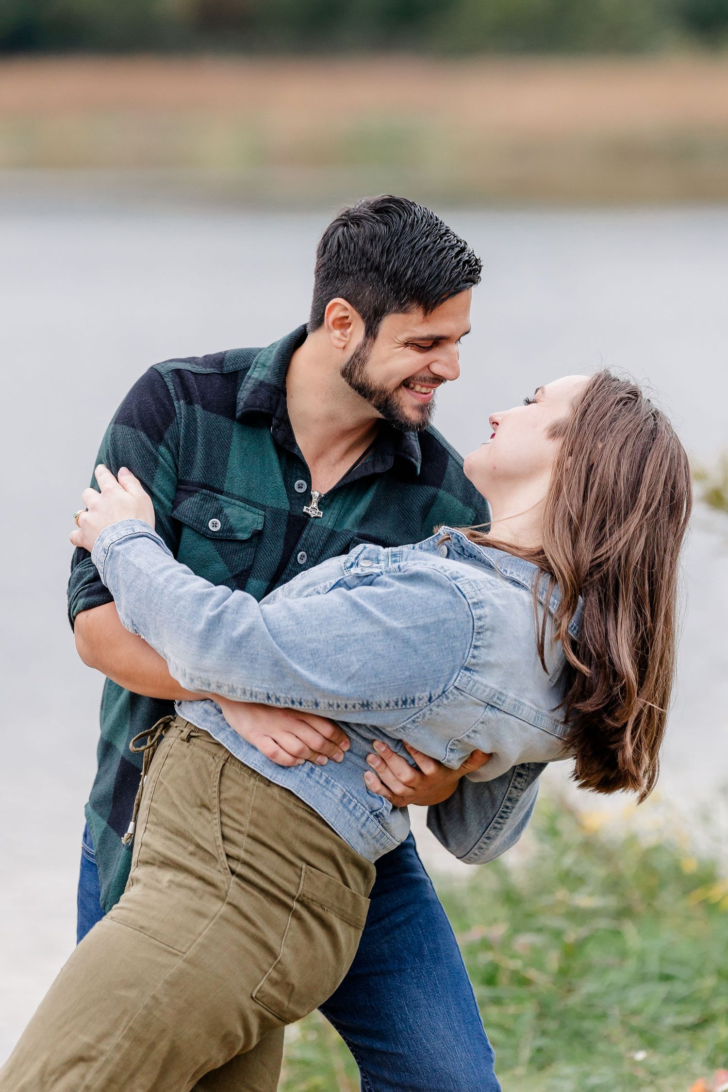 Man in a green plaid shirt dips a laughing woman in a denim jacket in a playful pose beside a lake during an engagement shoot.