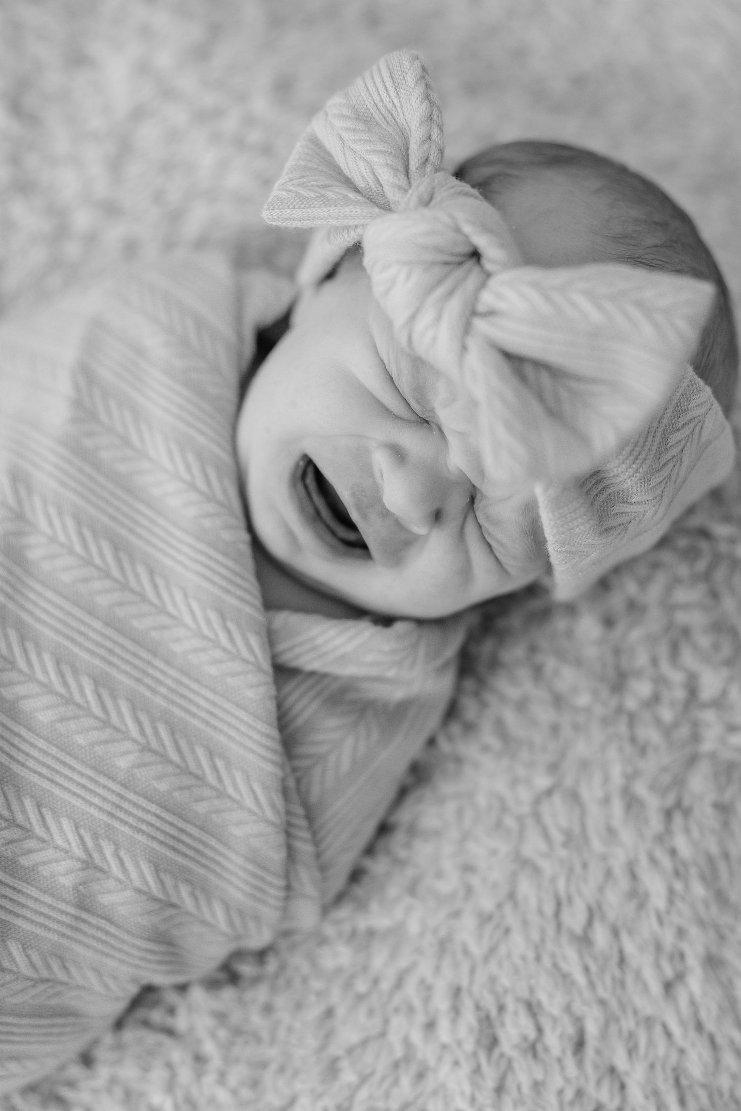 Black and white photo of a crying newborn baby girl swaddled in a blanket wearing a bow headband.