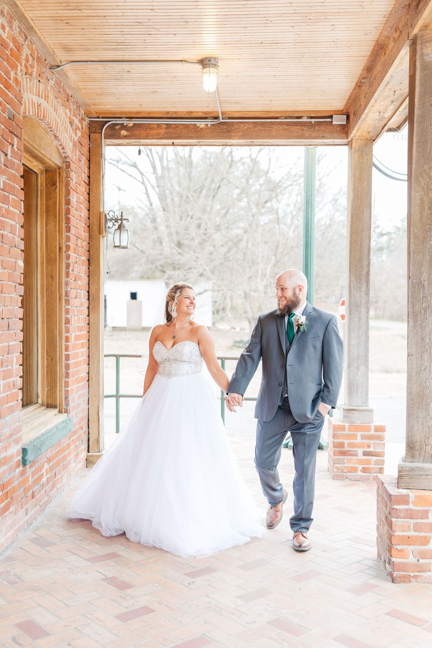 Newlyweds walking hand in hand on a rustic covered porch with brick columns.