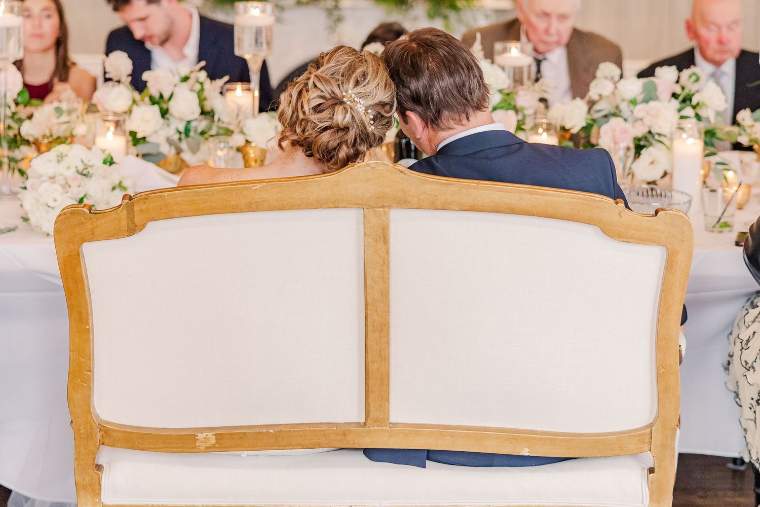 Couple seated on white upholstered loveseat at wedding reception.