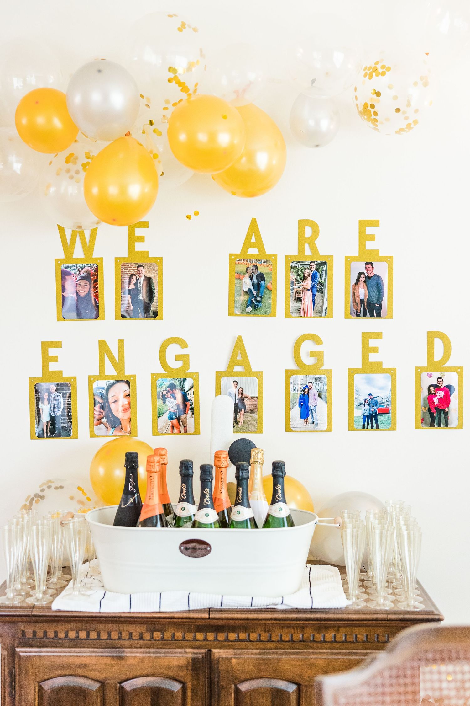 Yellow and white balloon display with engagement photos and champagne bottles on wooden sideboard.