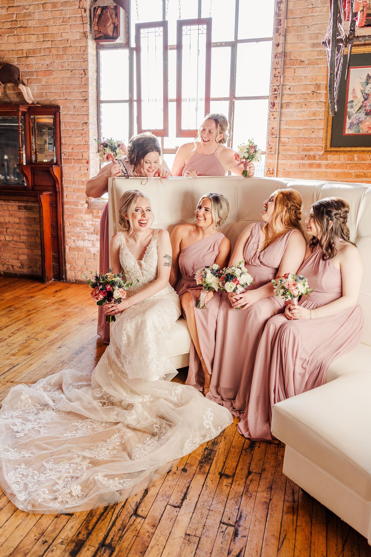 Bridal party in mauve dresses sharing laughs on white couch in rustic brick venue.