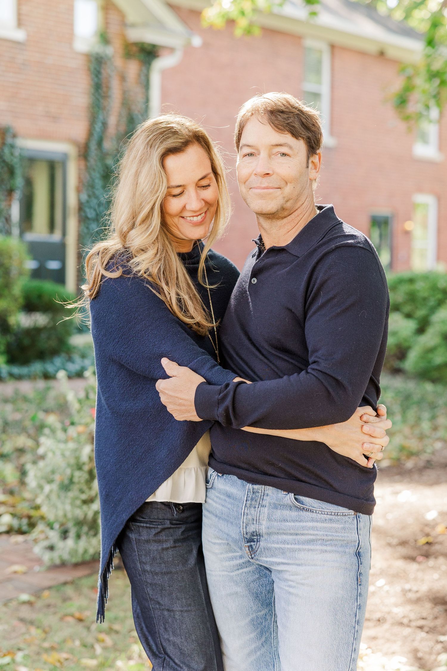 A couple in matching navy sweaters embraces outdoors in front of a brick building.