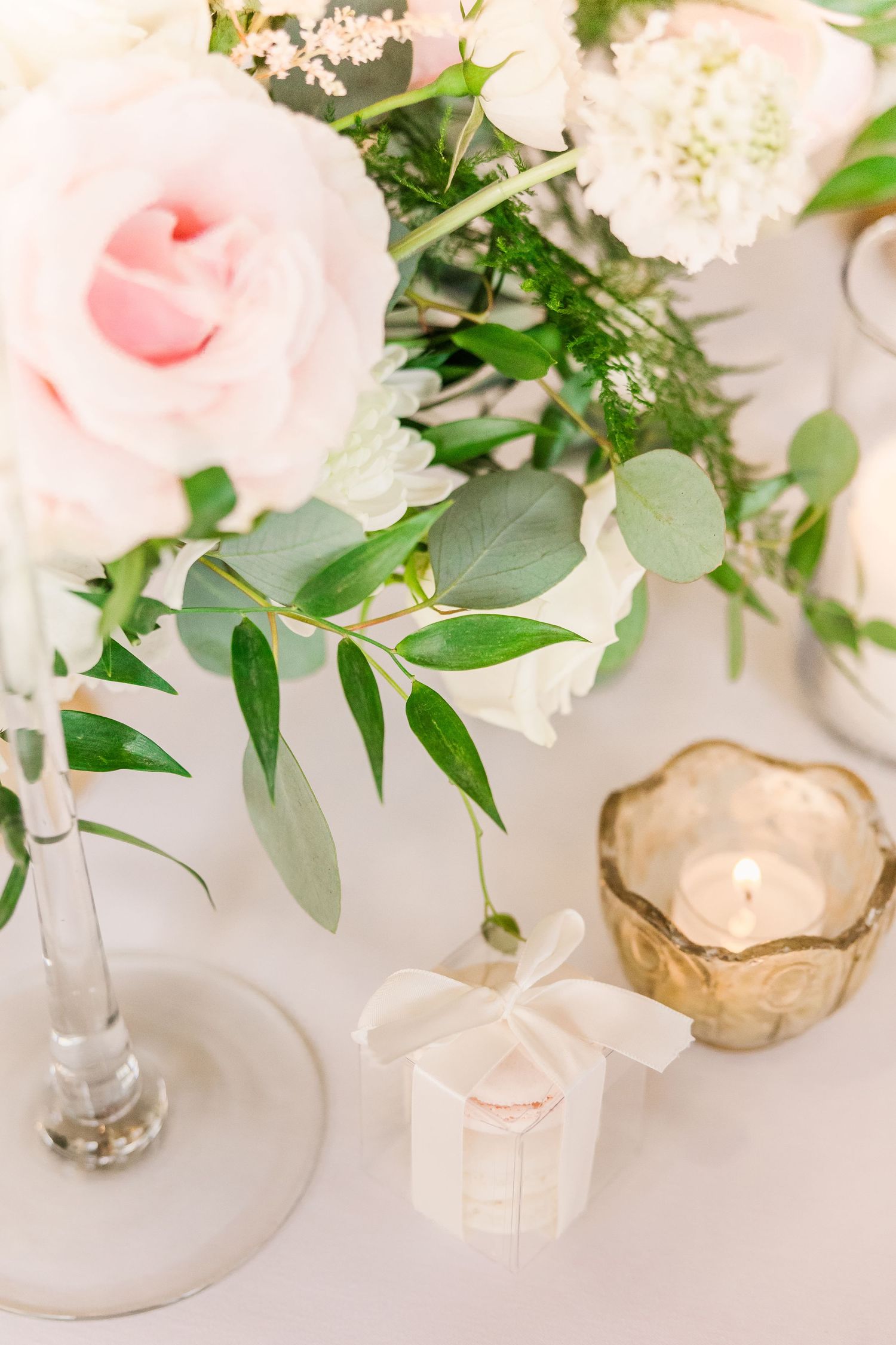 Soft pink roses and white florals with eucalyptus leaves arranged beside gold votives and favor boxes.