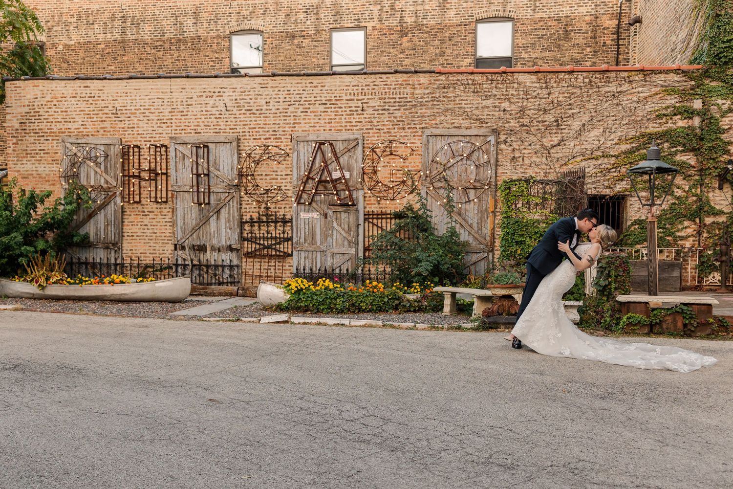 A couple embraces near an industrial brick wall with decorative iron gates and spring flowers.
