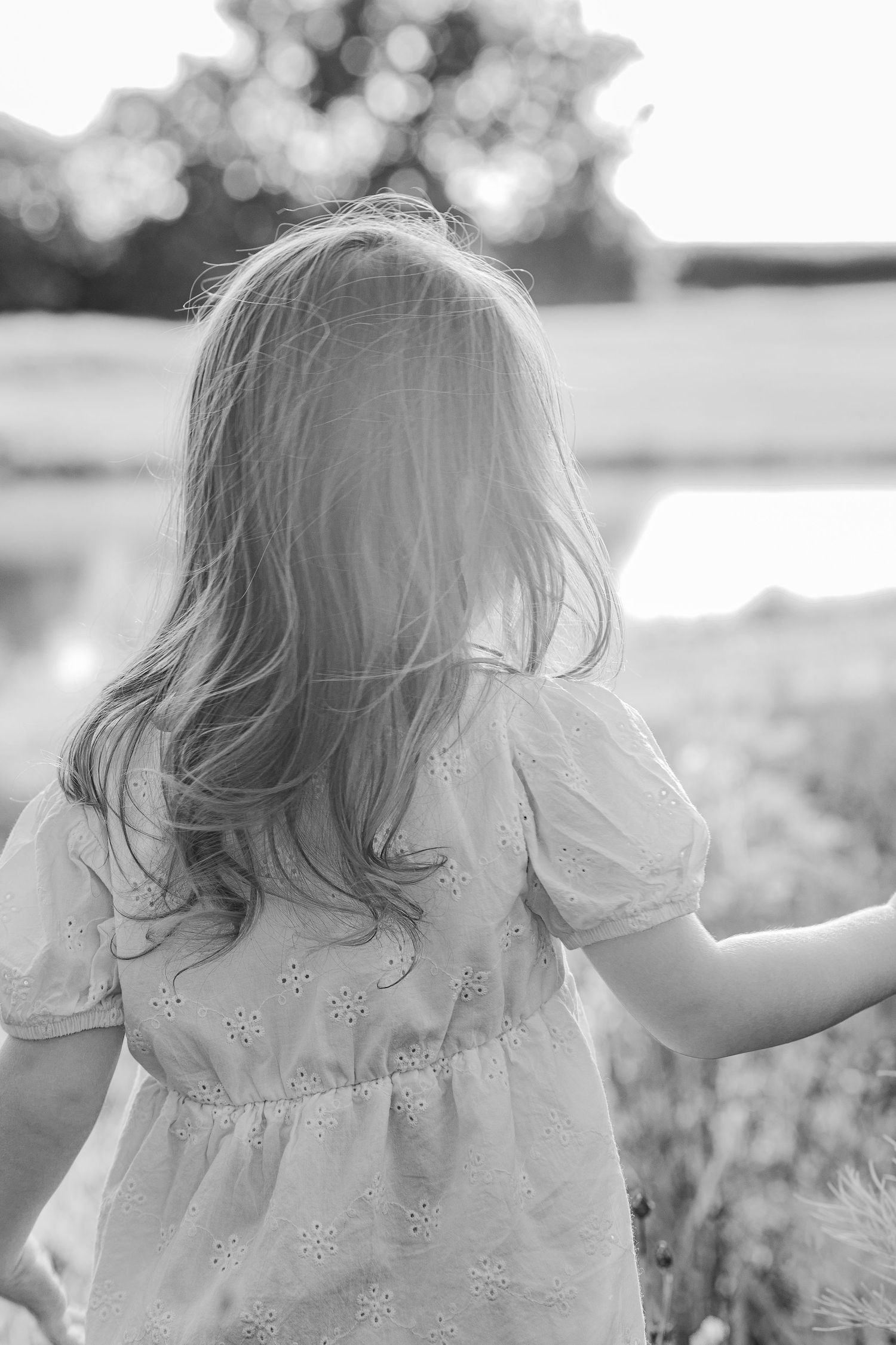 Black and white portrait of a young child with flowing hair in a floral dress standing in a sunlit field.
