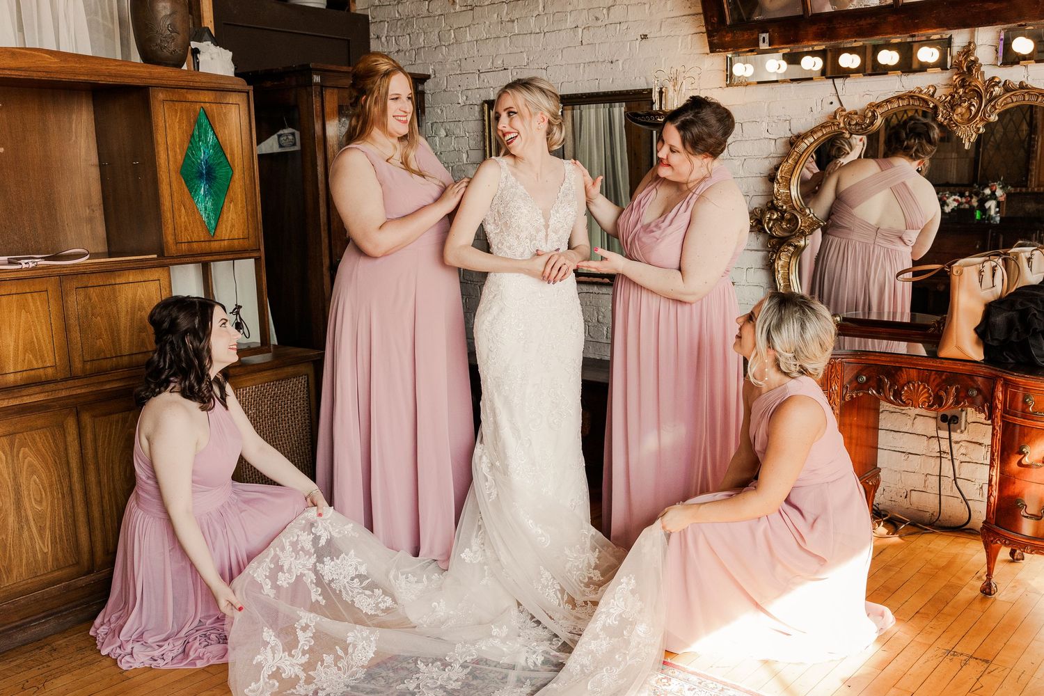 Wedding party in blush pink dresses helping adjust the bride's lace gown in rustic venue.