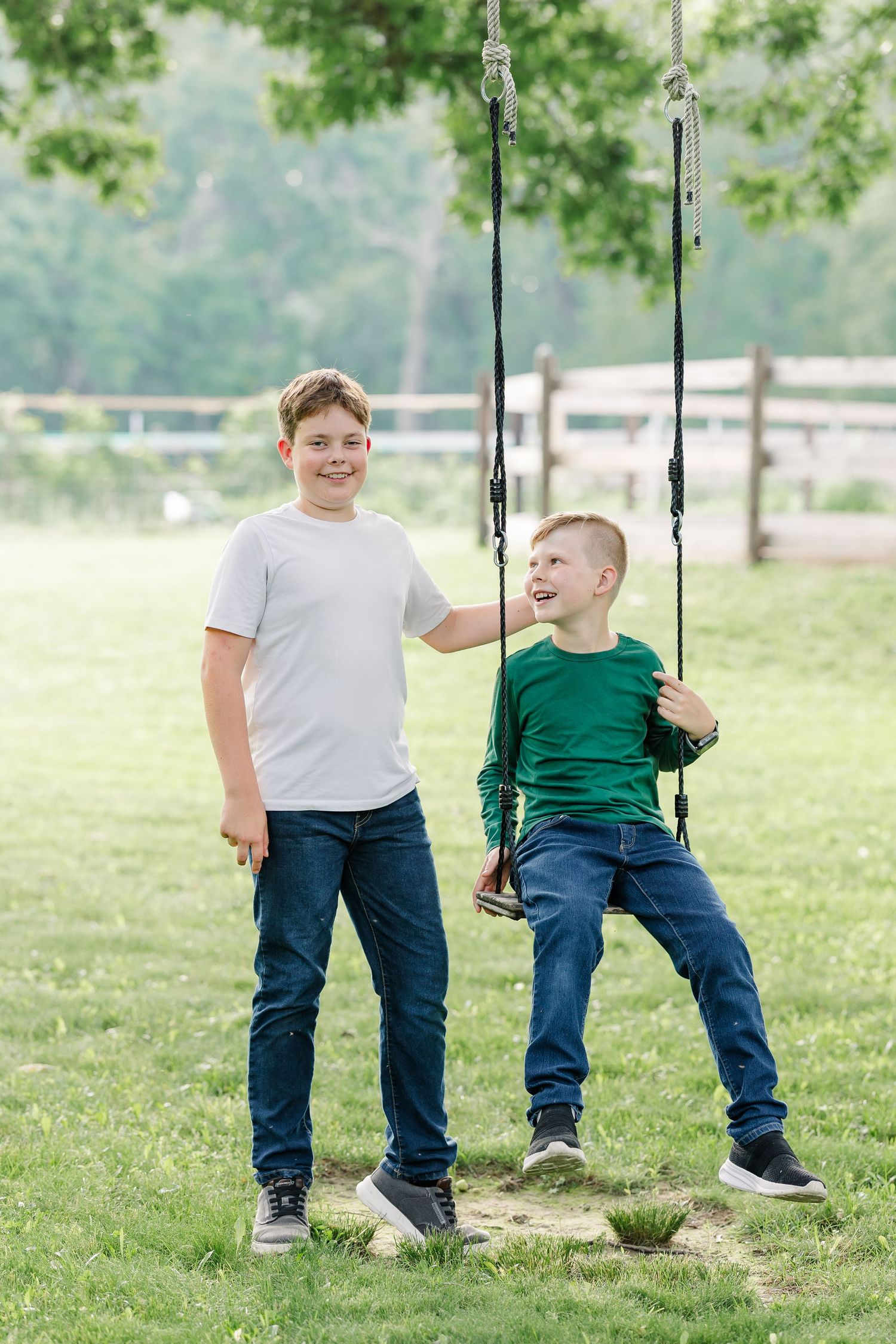 Two boys laughing and playing on a rope swing in a lush green park on a sunny day.