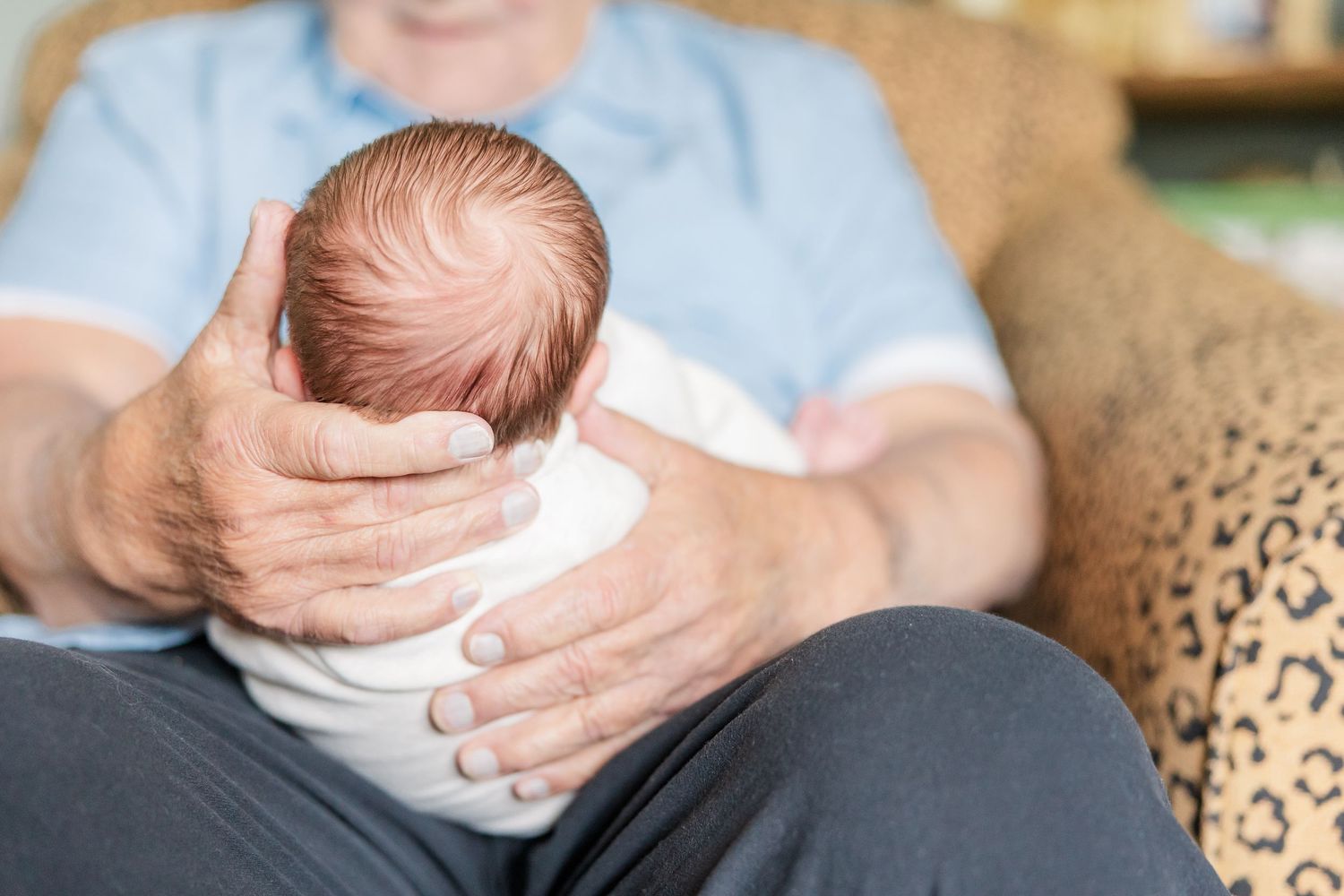 Grandparent gently cradling newborn baby's head while seated in leopard print chair.
