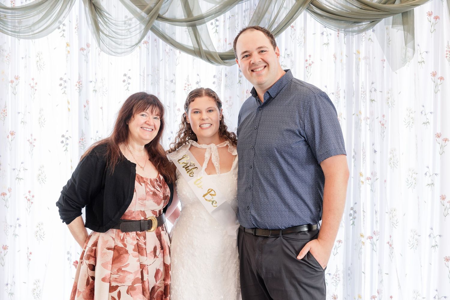 Three people smiling together in front of a floral curtain backdrop at a bridal shower celebration.
