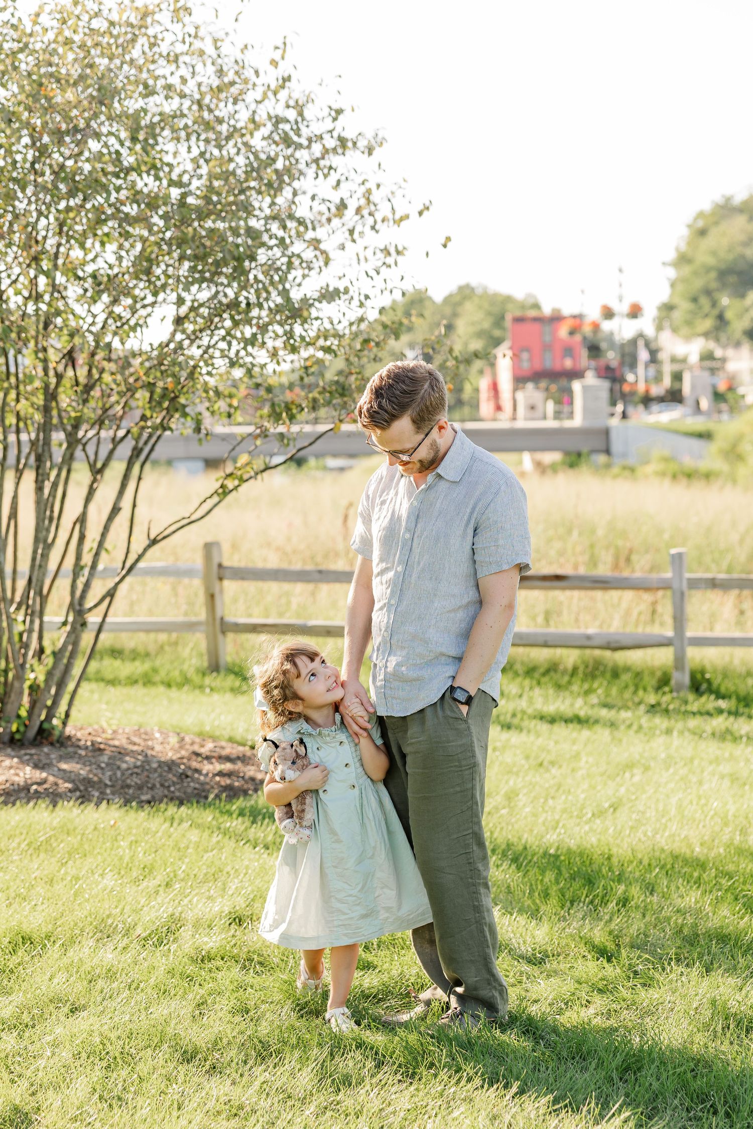 A father smiles down at his young daughter in a mint dress as they stand together on green grass outdoors.