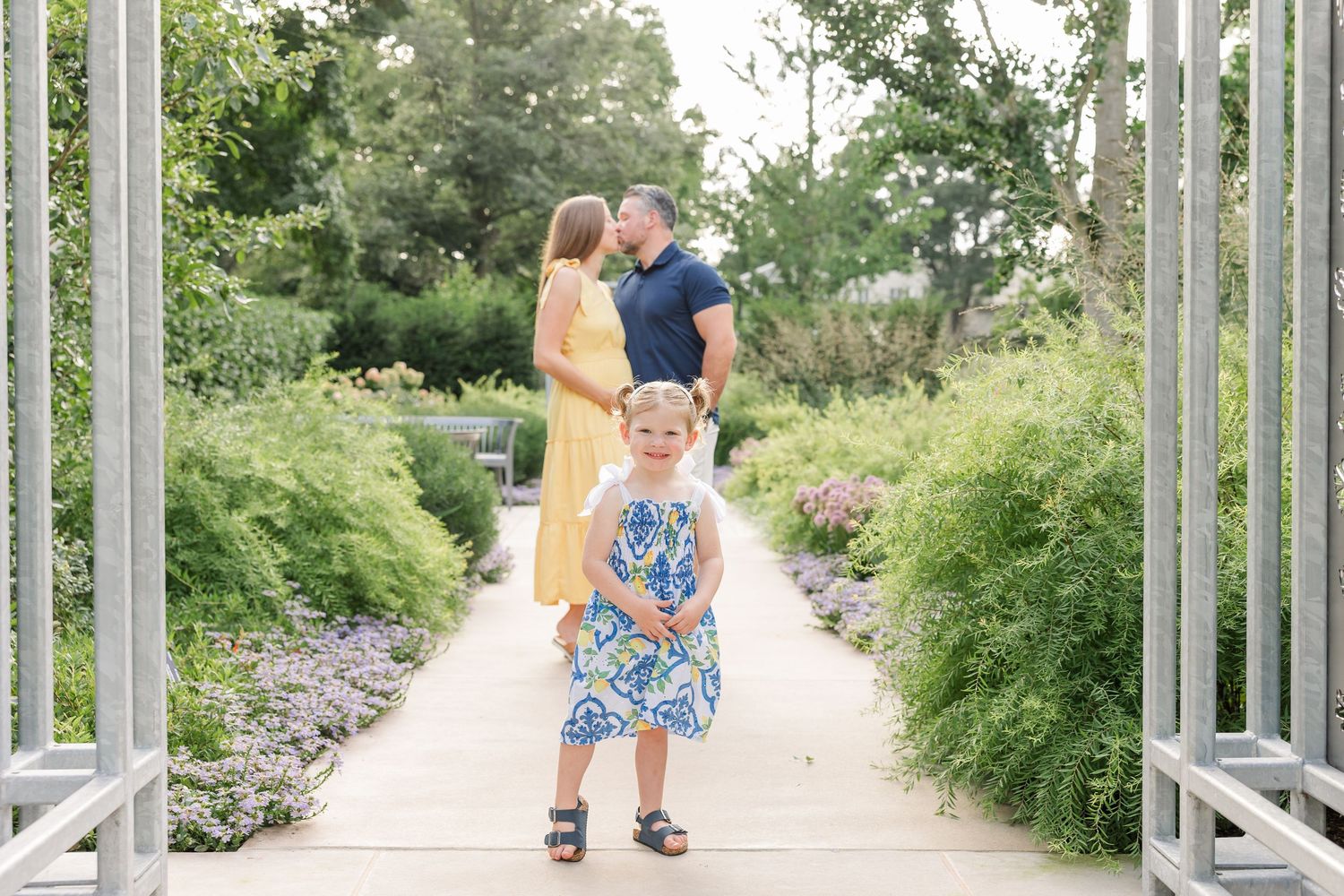 A young girl in a floral dress stands on a garden path while her parents share a romantic moment behind her.