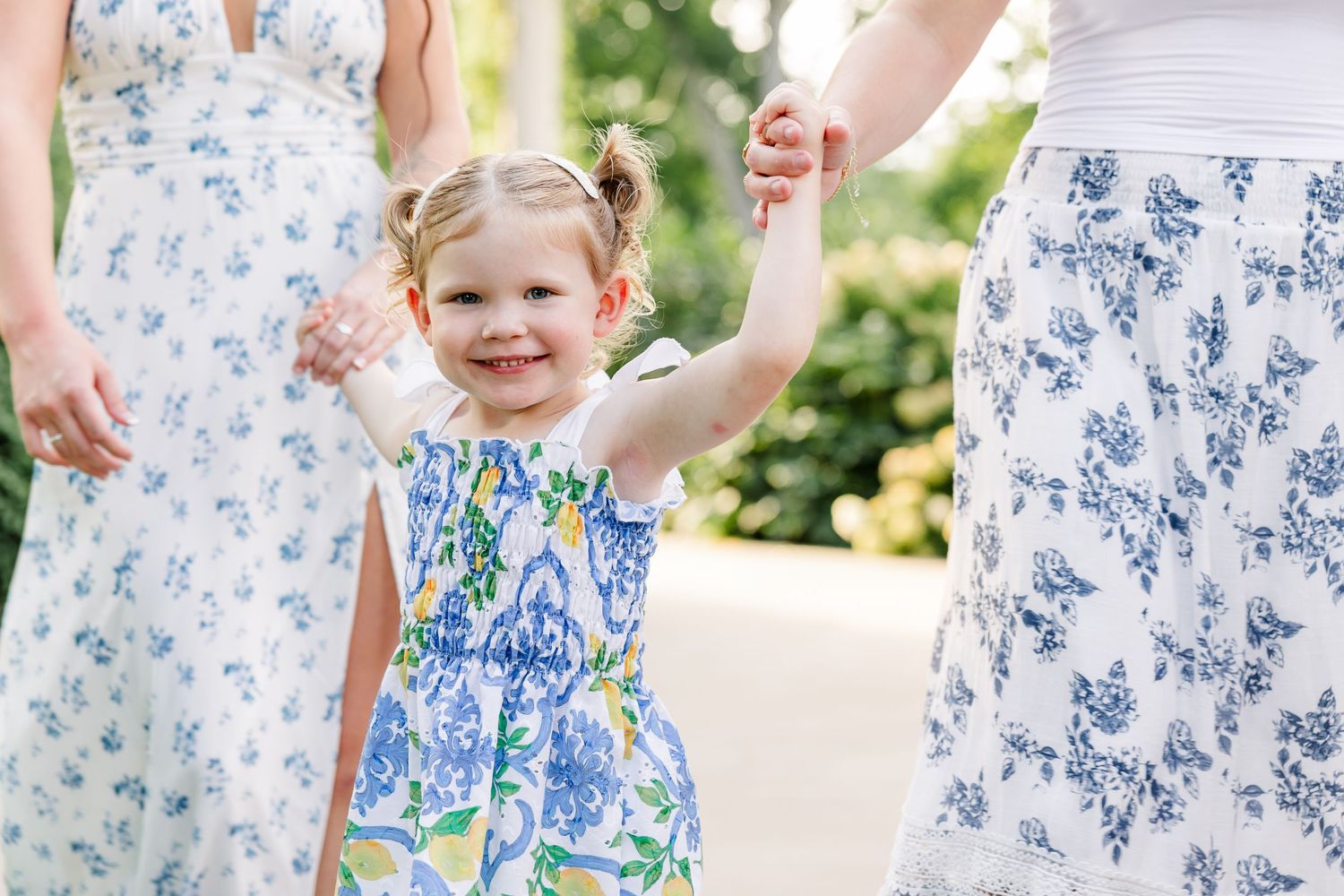 Smiling toddler girl in blue floral dress holds hands with women in a sunny garden.