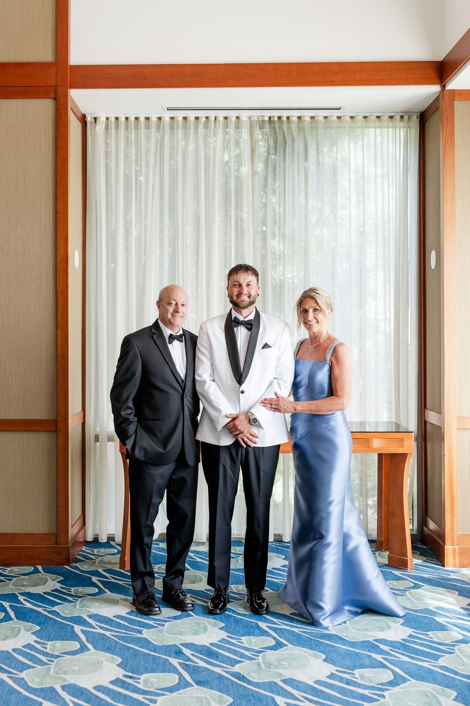 Groom in white tuxedo jacket poses with parents in black tuxedo and blue gown before wedding.