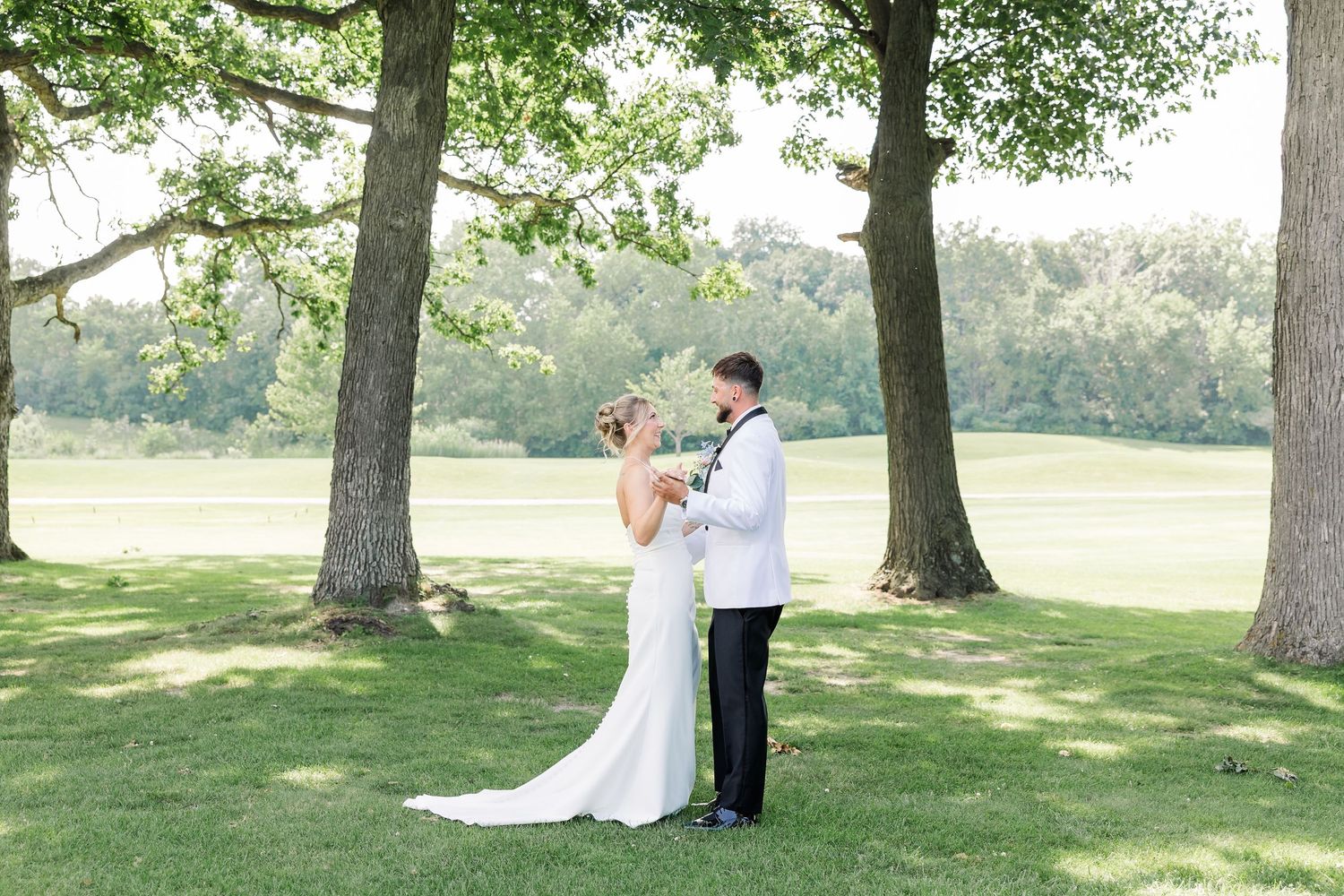 Bride and groom embrace during their first look under large trees on a scenic green golf course.